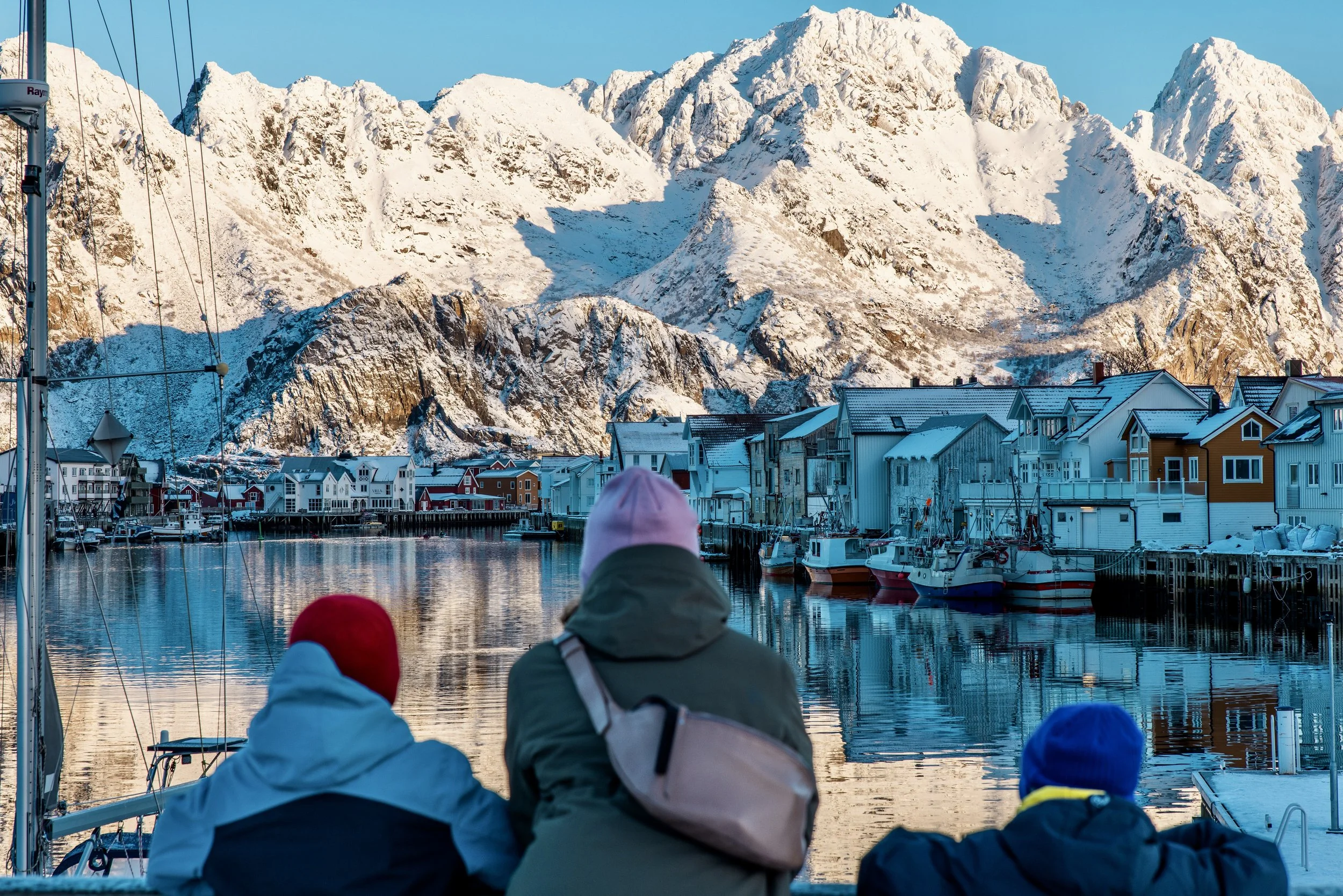 Three people with hoods and beanies sitting on a dock, overlooking a harbor with boats, colorful houses, and snow-capped mountains in the background.