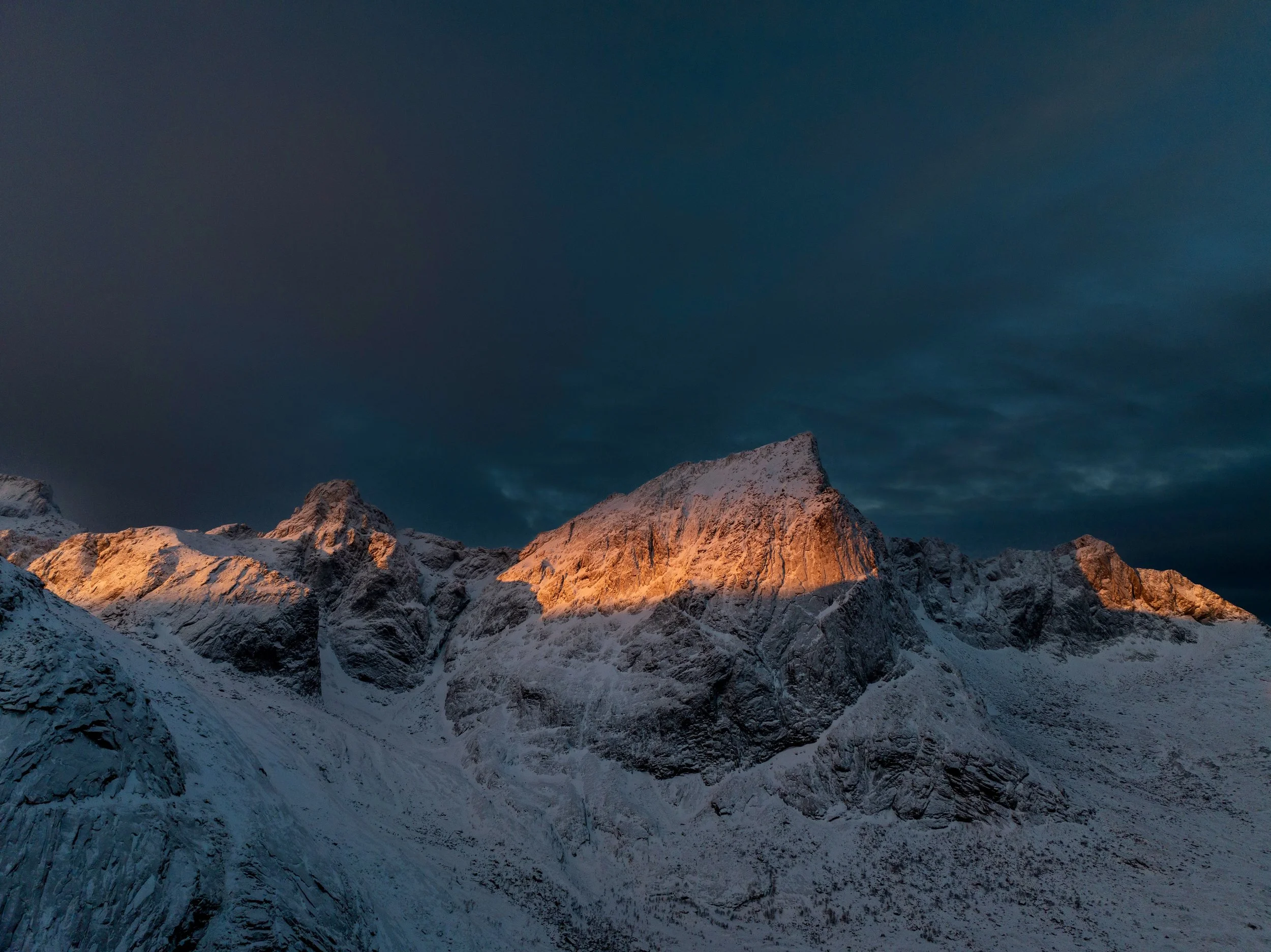 Snow-covered mountain peaks under dark sky, illuminated by warm sunlight at sunset.