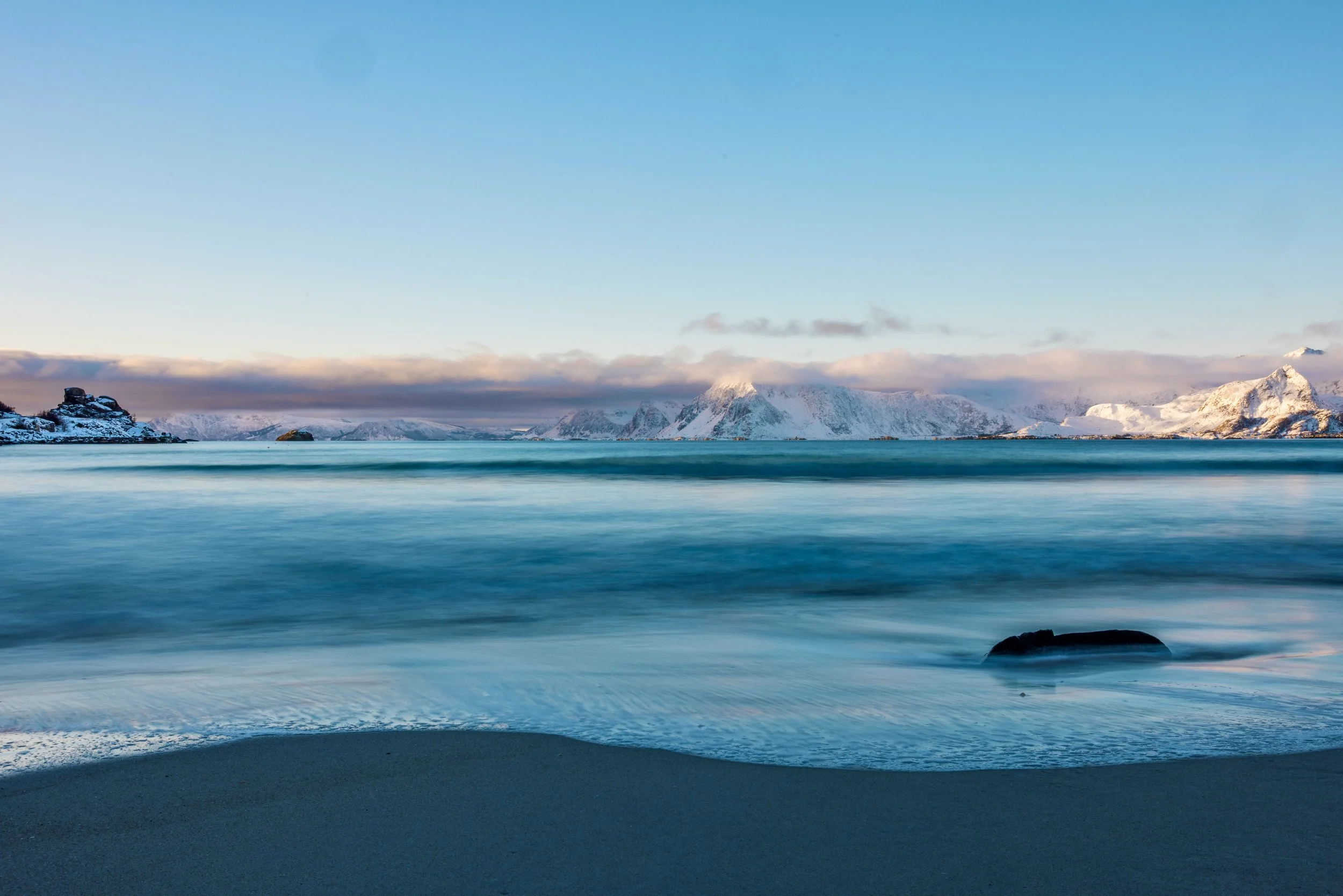 Snow-covered mountains across a calm ocean with sandy beach in foreground during sunset.