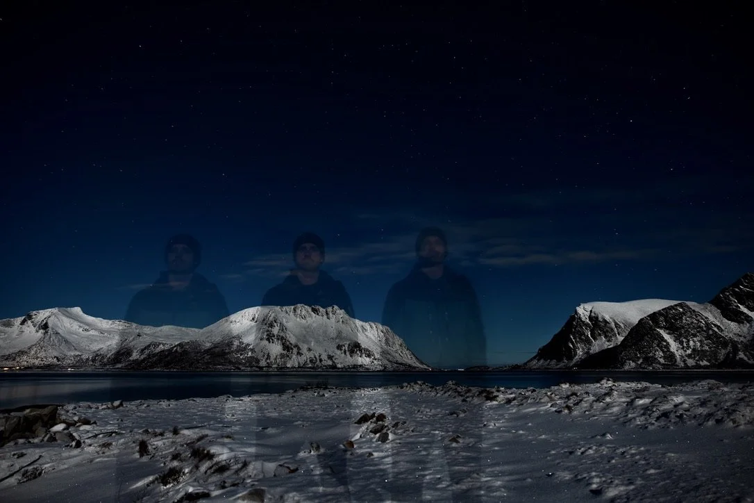 Nighttime snowy mountain landscape with a body of water, reflection of mountains, and faint stars in the sky, with three ghostly male figures superimposed in the sky.