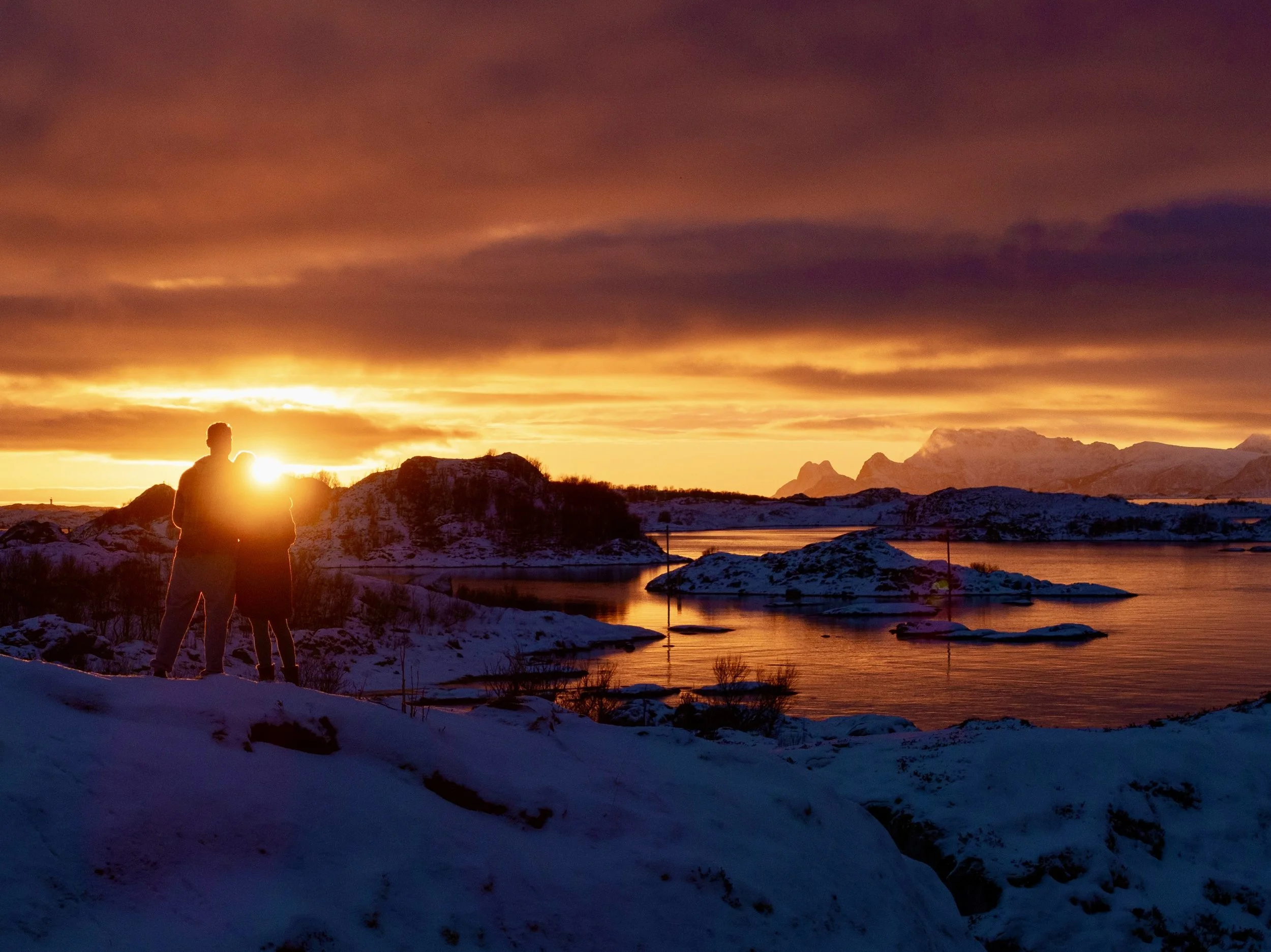 Silhouettes of a man and woman standing on snowy ground by a body of water during sunset, with mountains in the background and colorful sky.