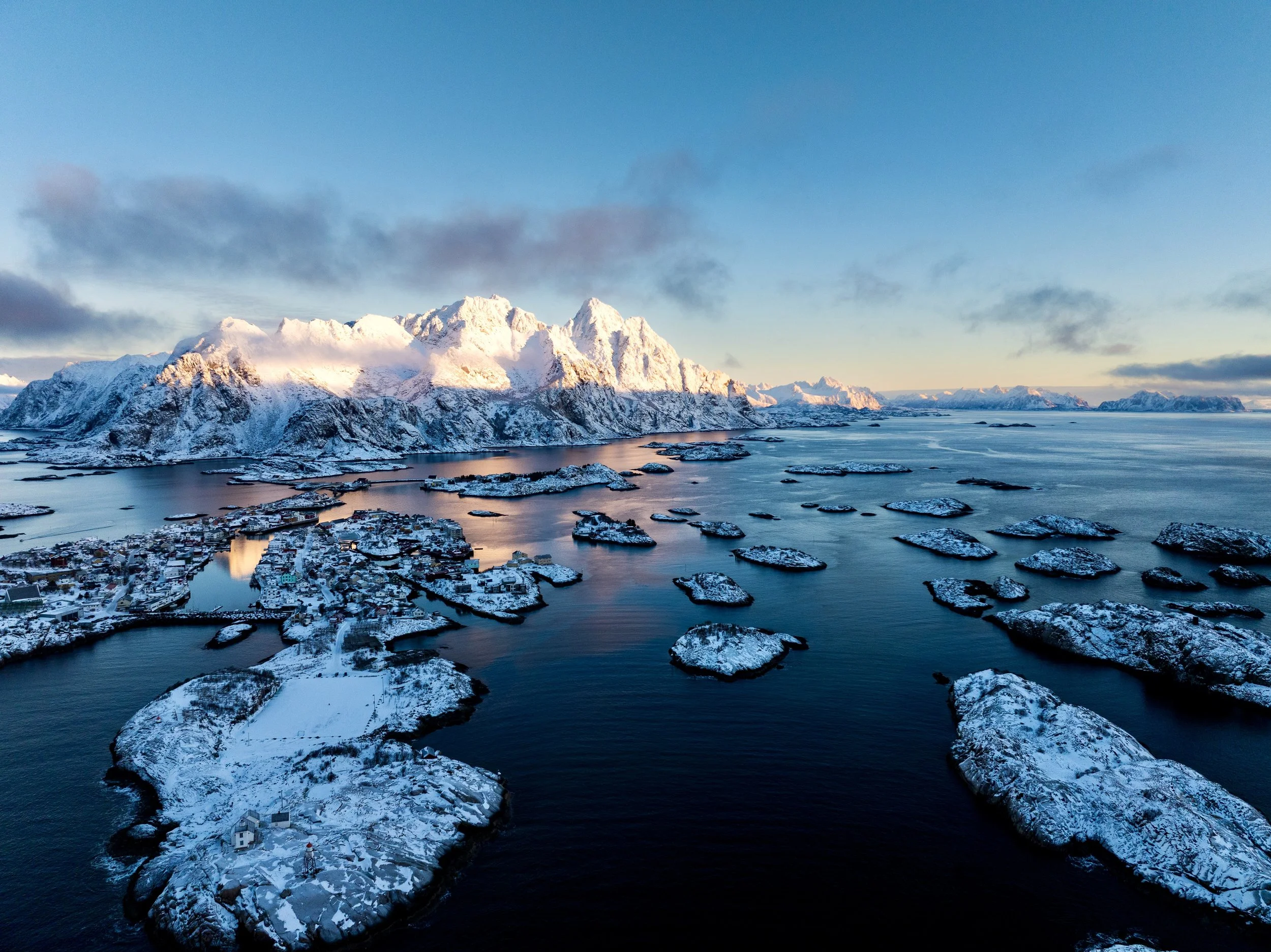 Snow-covered islands and a coastal town with mountains in the background during winter, with a partly cloudy sky.