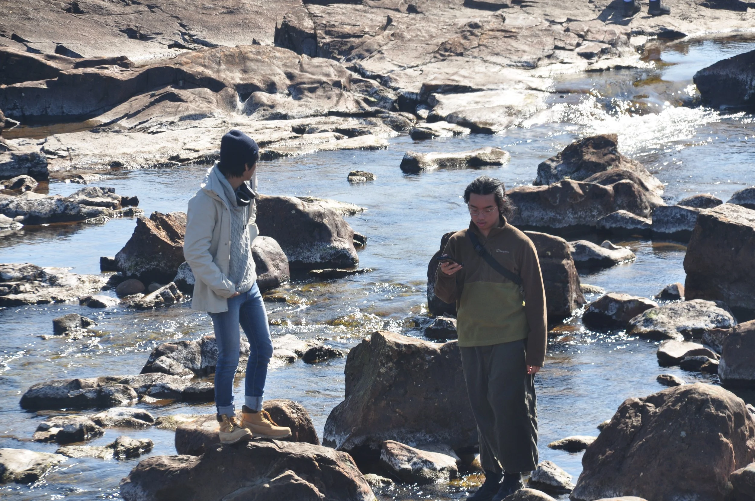 Two young men standing on rocks in a shallow river, one looking at his phone, surrounded by large rocks and flowing water.