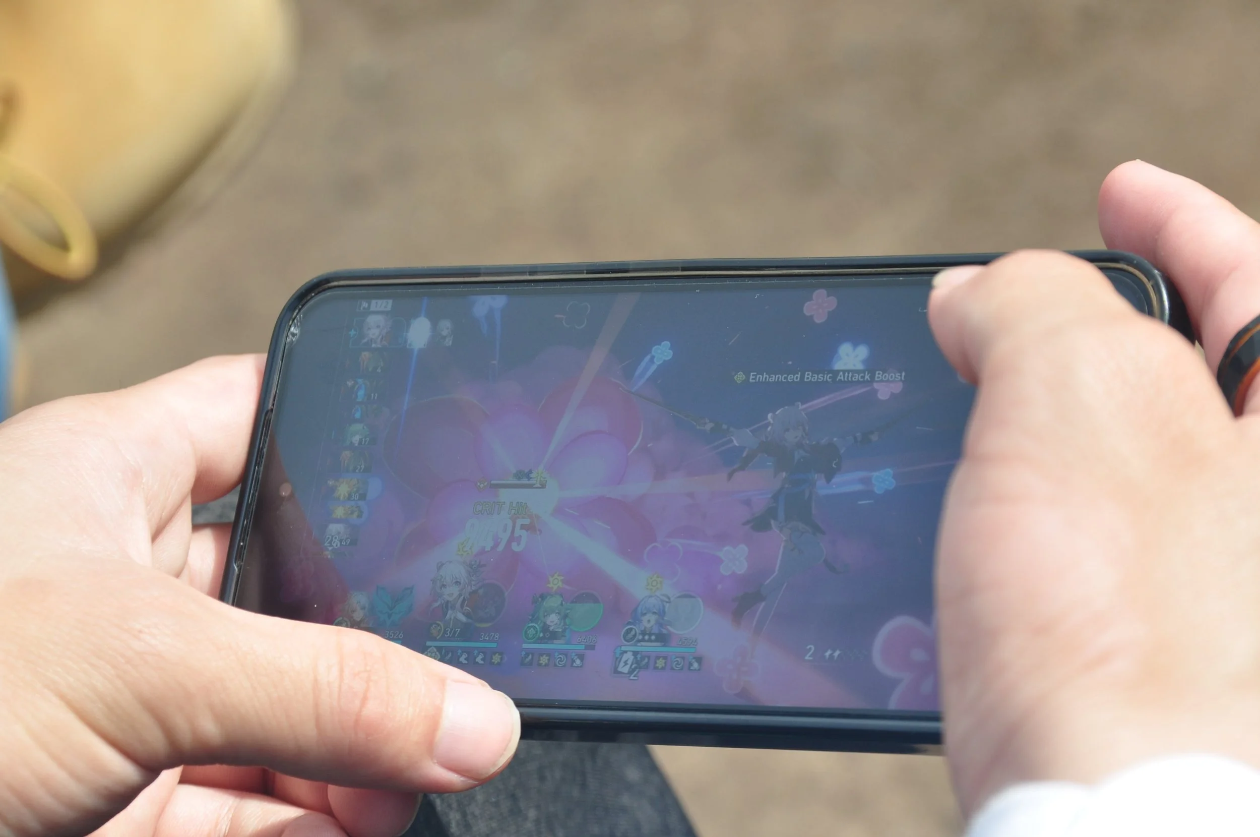 A person holding a smartphone playing a Japanese-themed anime-style game, with character icons and attack effects visible on the screen.