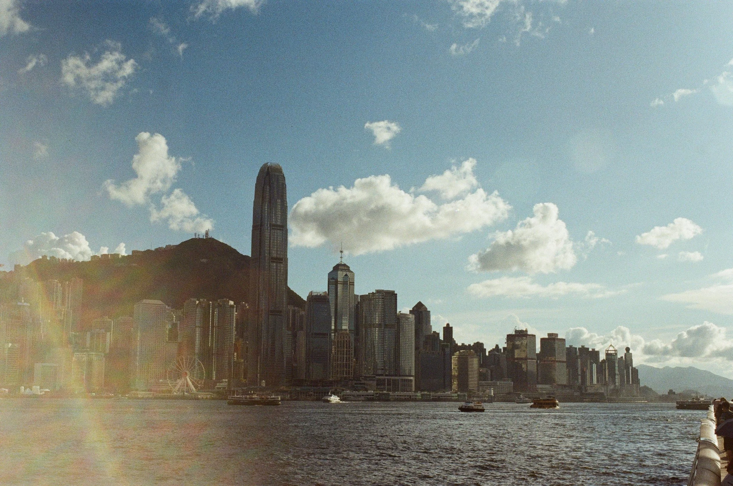 Hong Kong skyline with tall skyscrapers along Victoria Harbour, including the International Finance Centre, under a partly cloudy sky with sunlight, boats on the water, and mountains in the background.