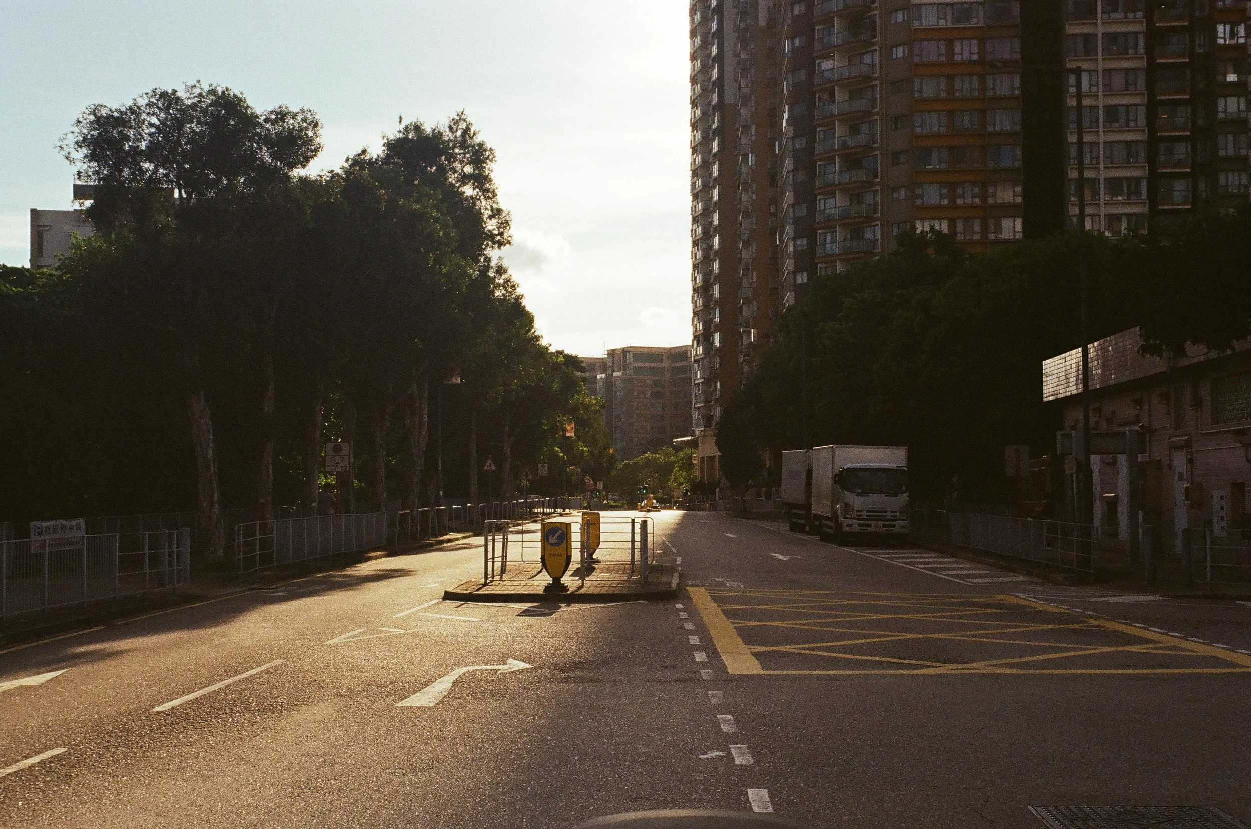 Empty city street with trees on the left and tall apartment buildings on the right, with a delivery truck parked on the right side, and yellow traffic lines and barriers in the foreground, during sunset.