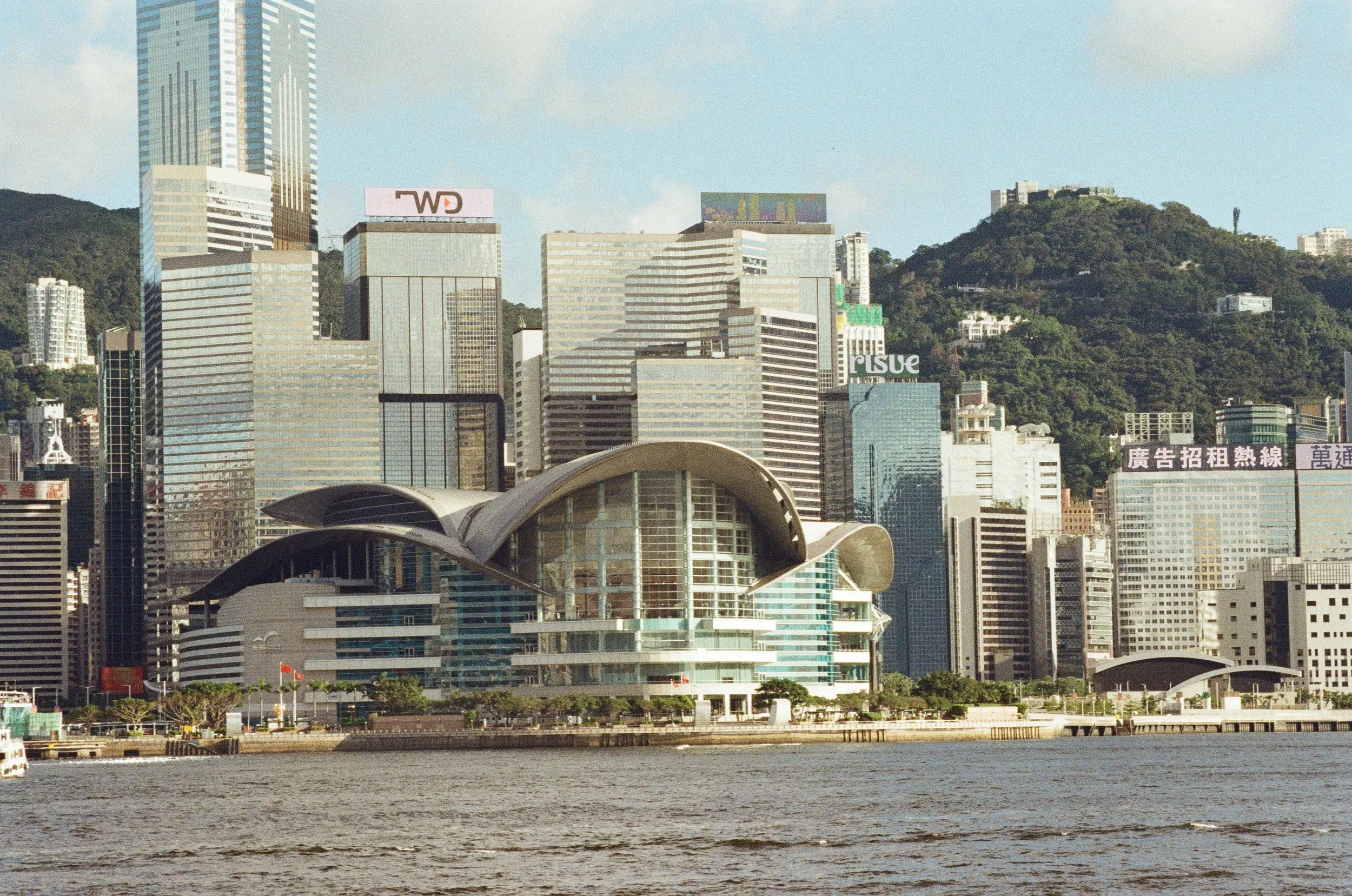 Photo of a city skyline with modern skyscrapers and a waterfront in the foreground, hills in the background, and cloudy sky.