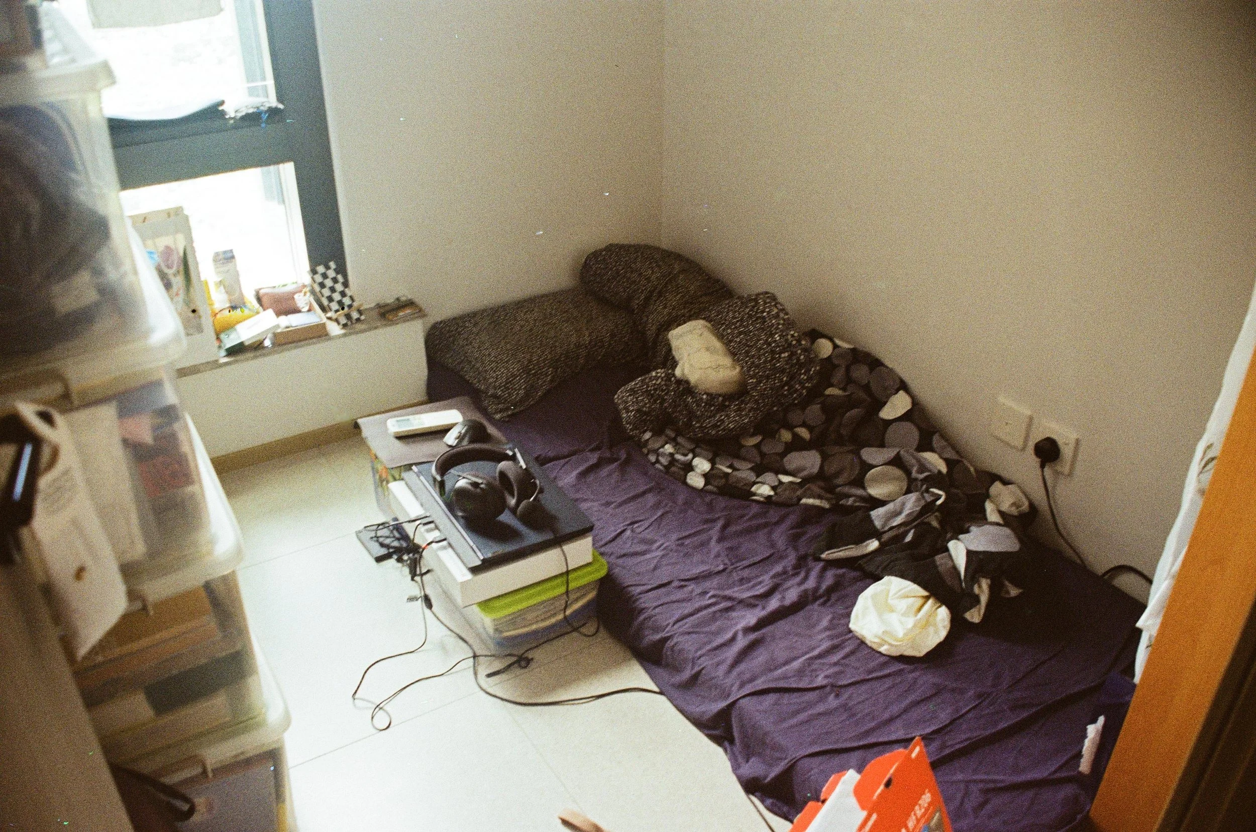 A small, cluttered bedroom with a mattress on the floor covered with dark and patterned sheets, and four pillows, two with black and white patterns and one white fabric pillow. Next to the mattress, there's a pile of electronic devices, including hea