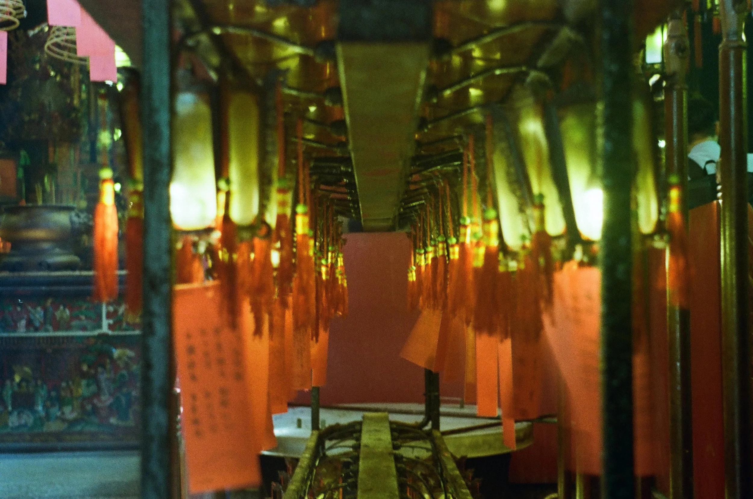 Rows of hanging lanterns and banners in a temple with a glass-like reflective ceiling or floor, illuminated by warm yellow and green lights.