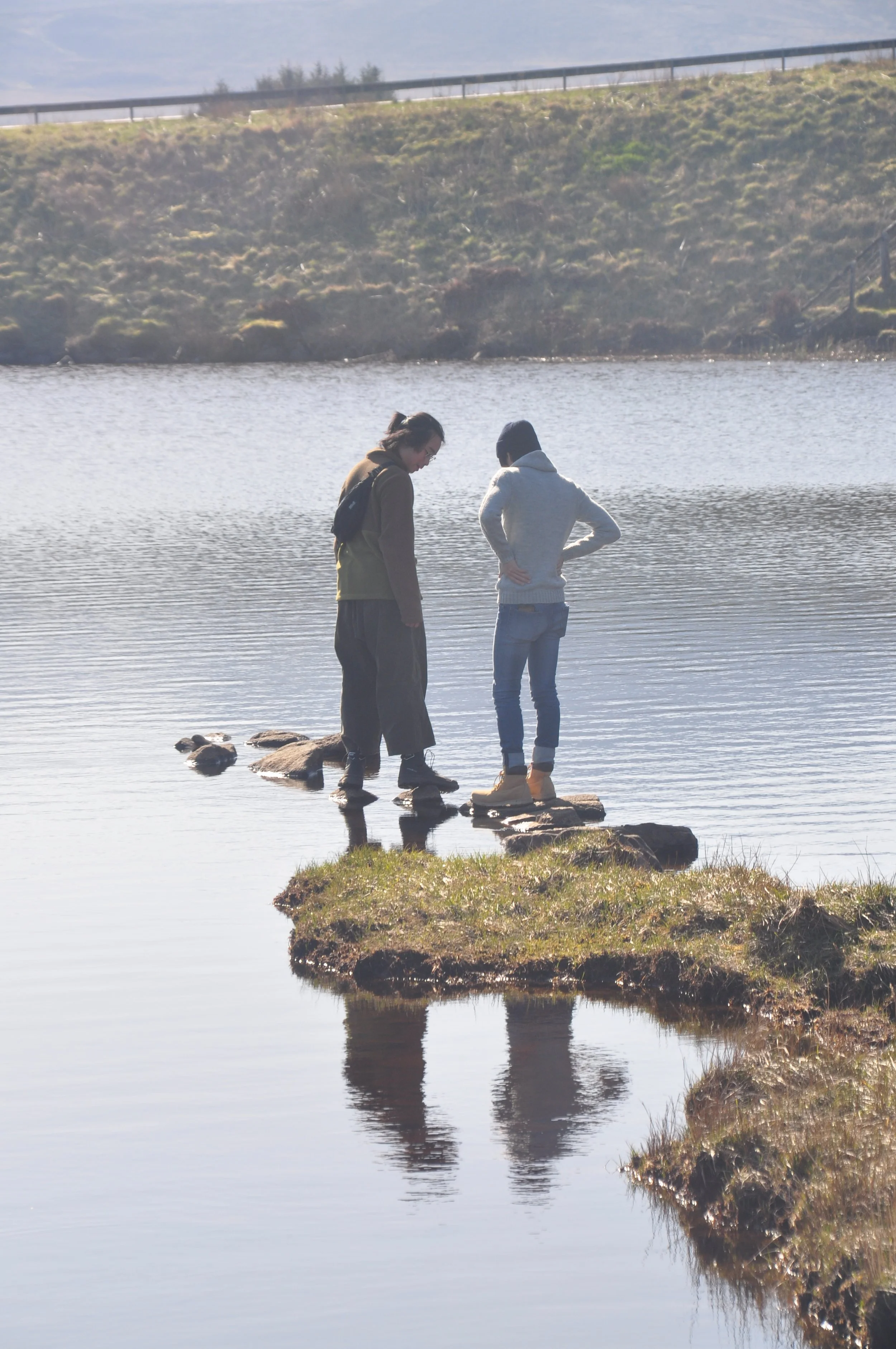 Two people standing on rocks in a calm lake, looking at each other, with grassy patches on the shore and a hill in the background.