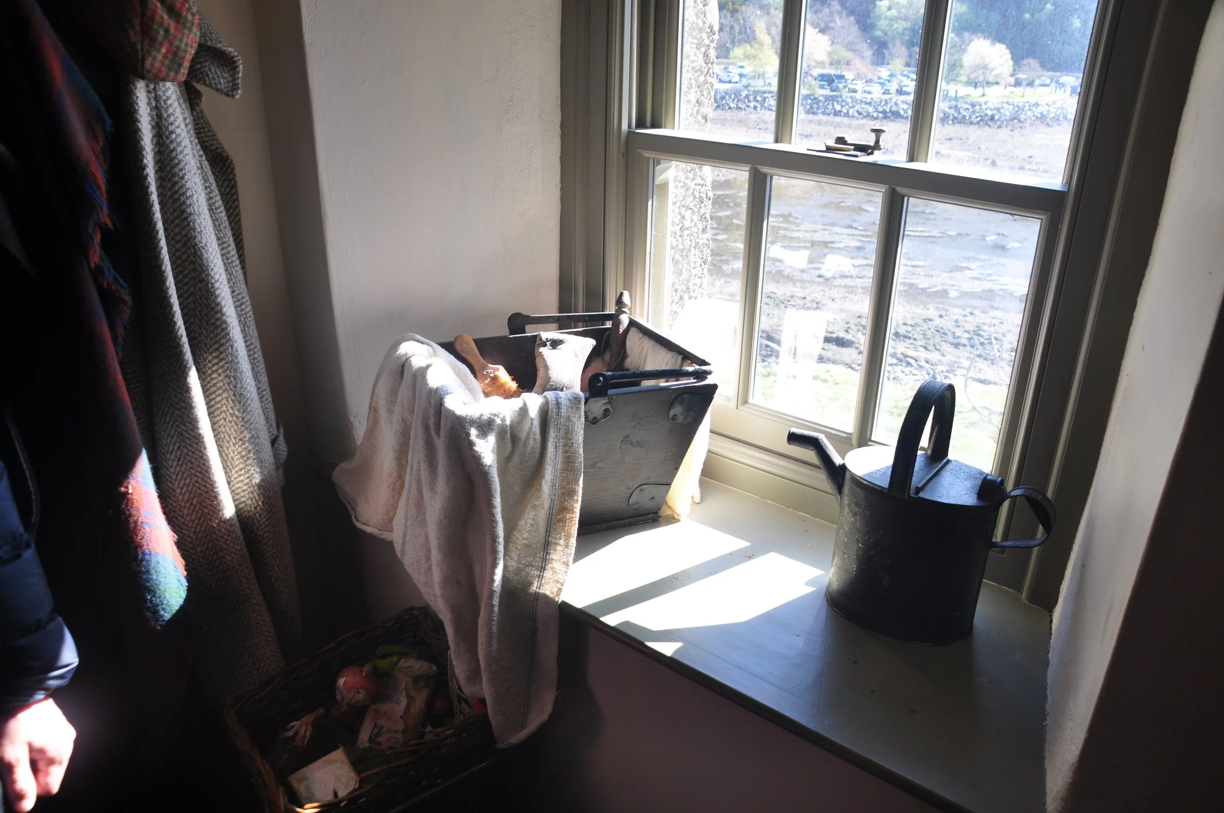 A windowsill with a black watering can, a wooden box with gardening tools and a towel, and a woven basket with gardening supplies, in a bright room with sunlight streaming through the window.