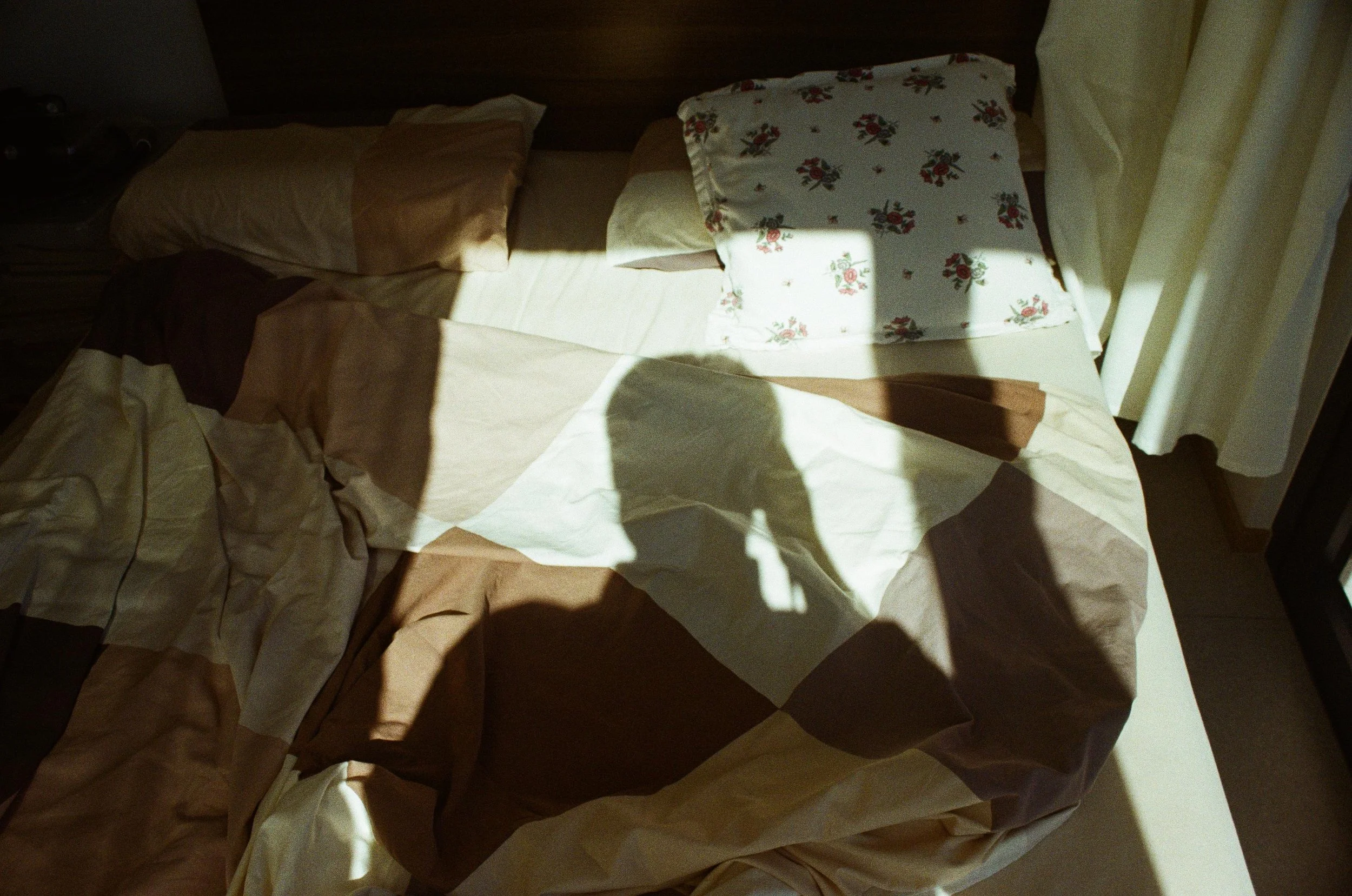 A bed with crumpled sheets, a floral pillow, and sunlight casting shadows, including a shadow of a person taking the photo.