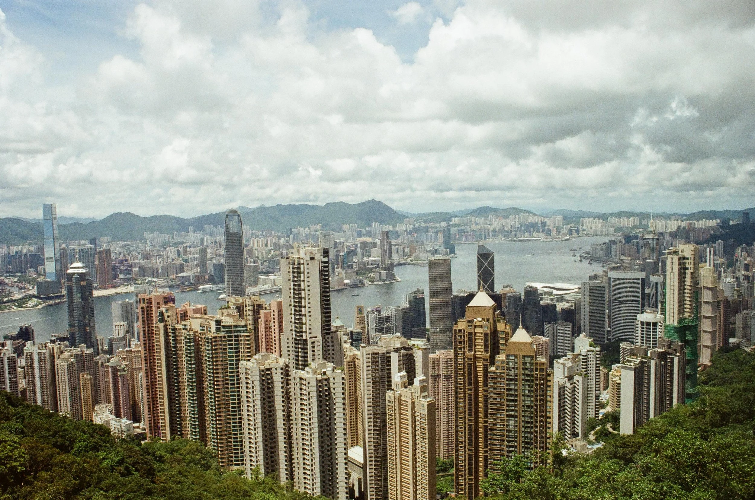 A city skyline with numerous high-rise buildings, a river in the middle, and mountains in the background under a cloudy sky.