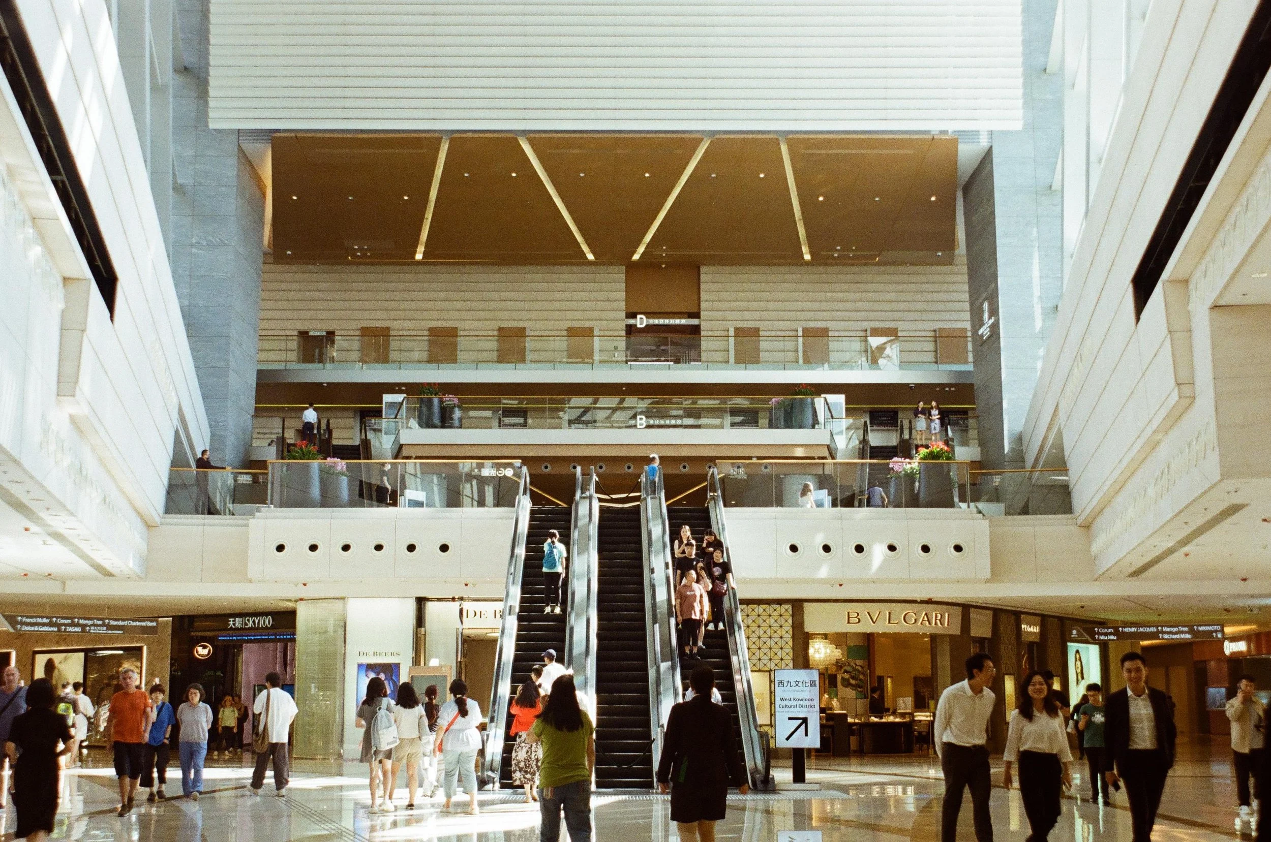 Interior of a shopping mall with people walking, an escalator in the center, and luxury stores like BVLGARI at the bottom.