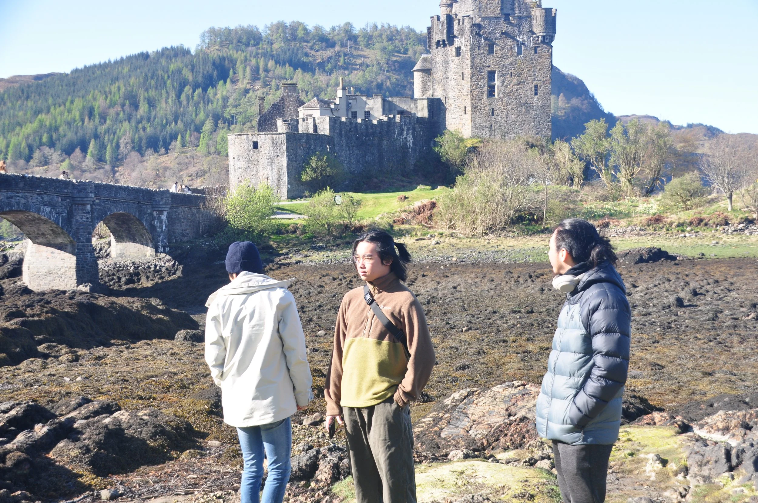 Three people standing outdoors near a rocky area with a castle on a hill in the background. One person is facing away wearing a beanie and white jacket, another is looking towards the camera wearing glasses and a brown jacket, and the third person is