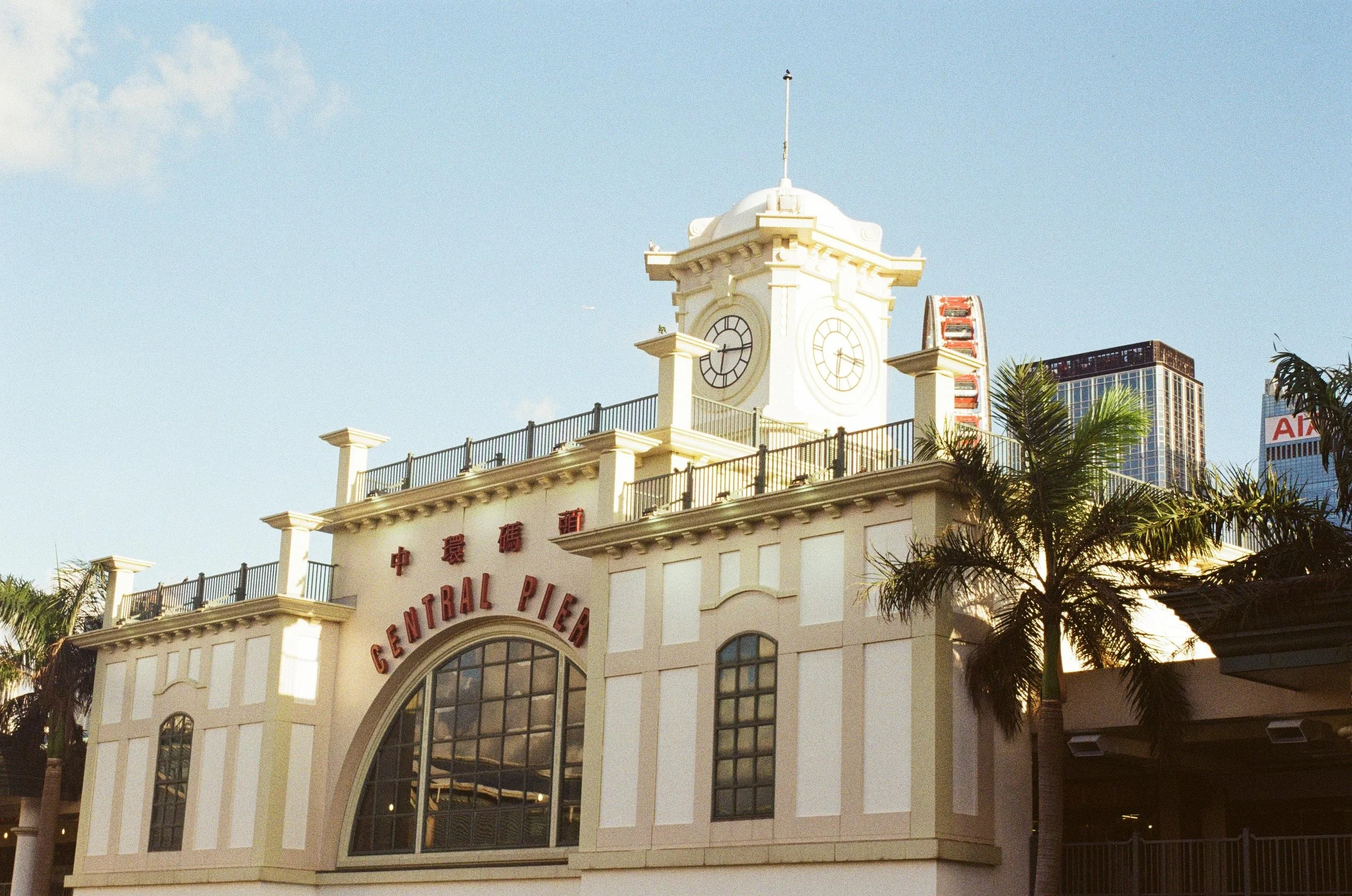 Front of Central Pier building with clock tower, palm trees, and modern high-rise buildings in the background, under a blue sky.