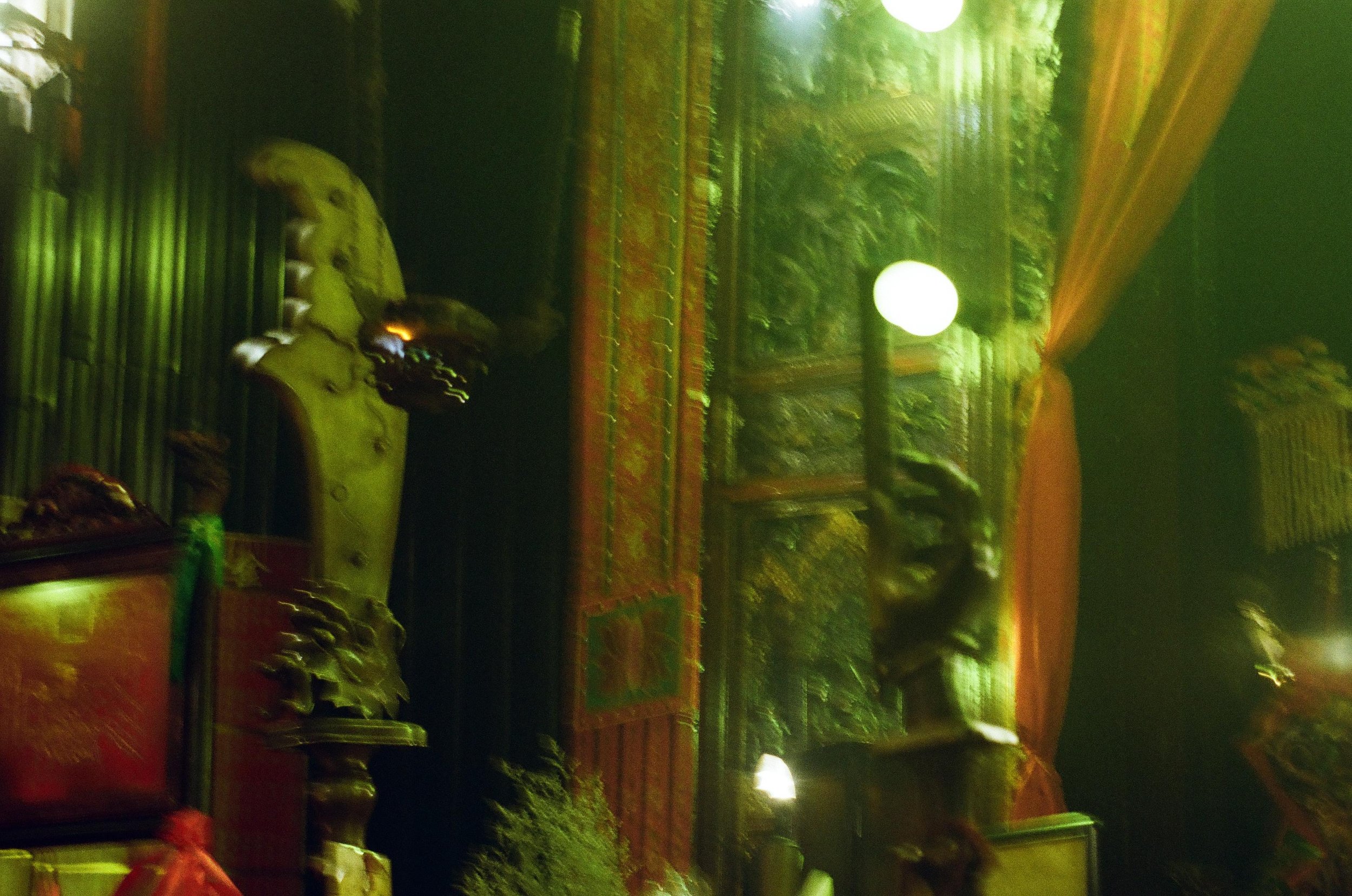 An interior of a traditional room with ornate wooden furniture, carved sculptures, and hanging drapes, illuminated by green lighting and round ceiling lights.