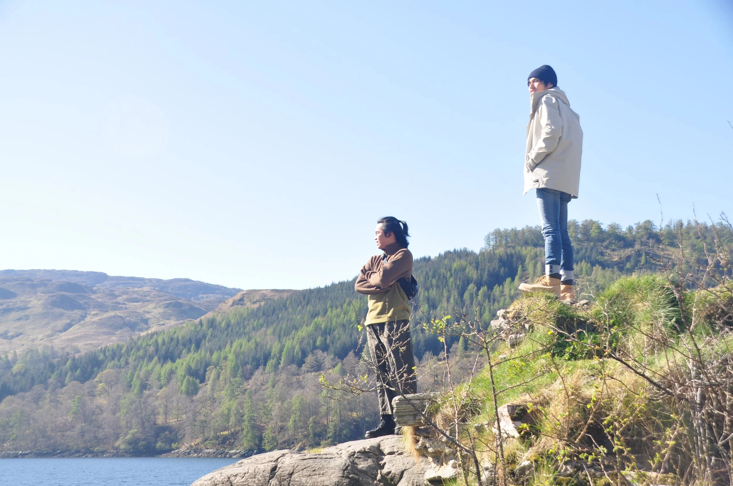 Two women standing on rocks near a lake, with lush green hills and mountains in the background, under a clear blue sky.