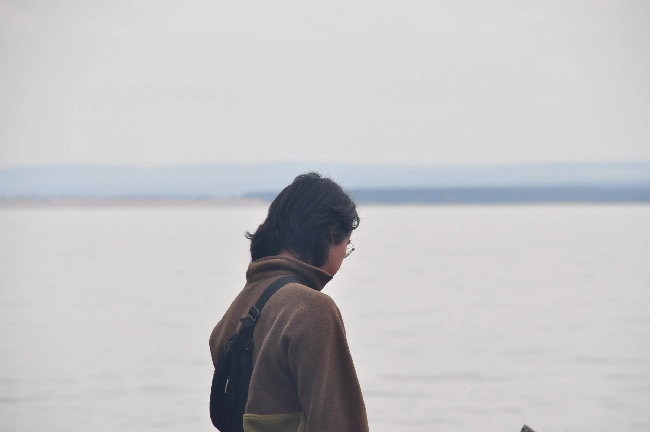 Person with dark hair, glasses, and a brown sweater, looking down by a calm body of water, with a cloudy sky and distant land in the background.