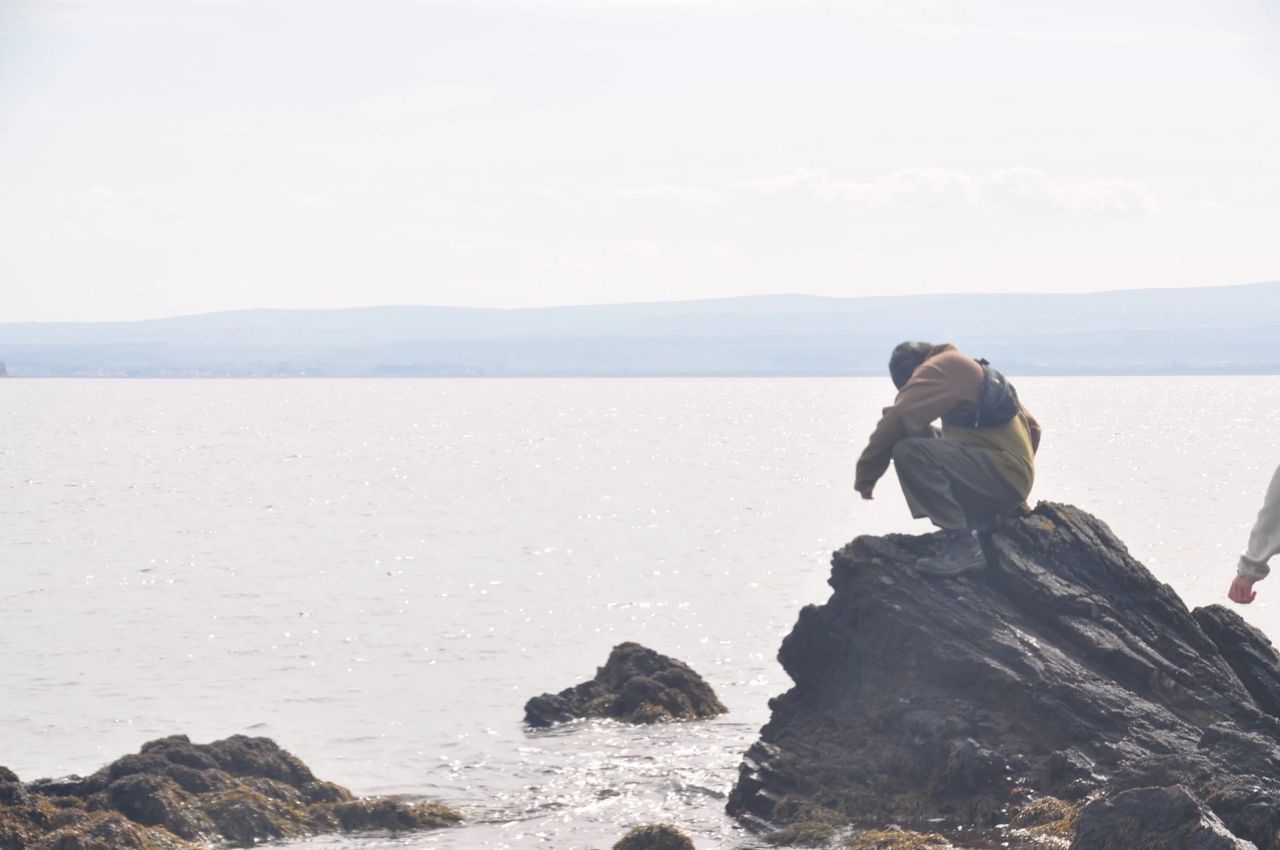Person sitting on a large rock by the water during daytime, with distant hills in the background.
