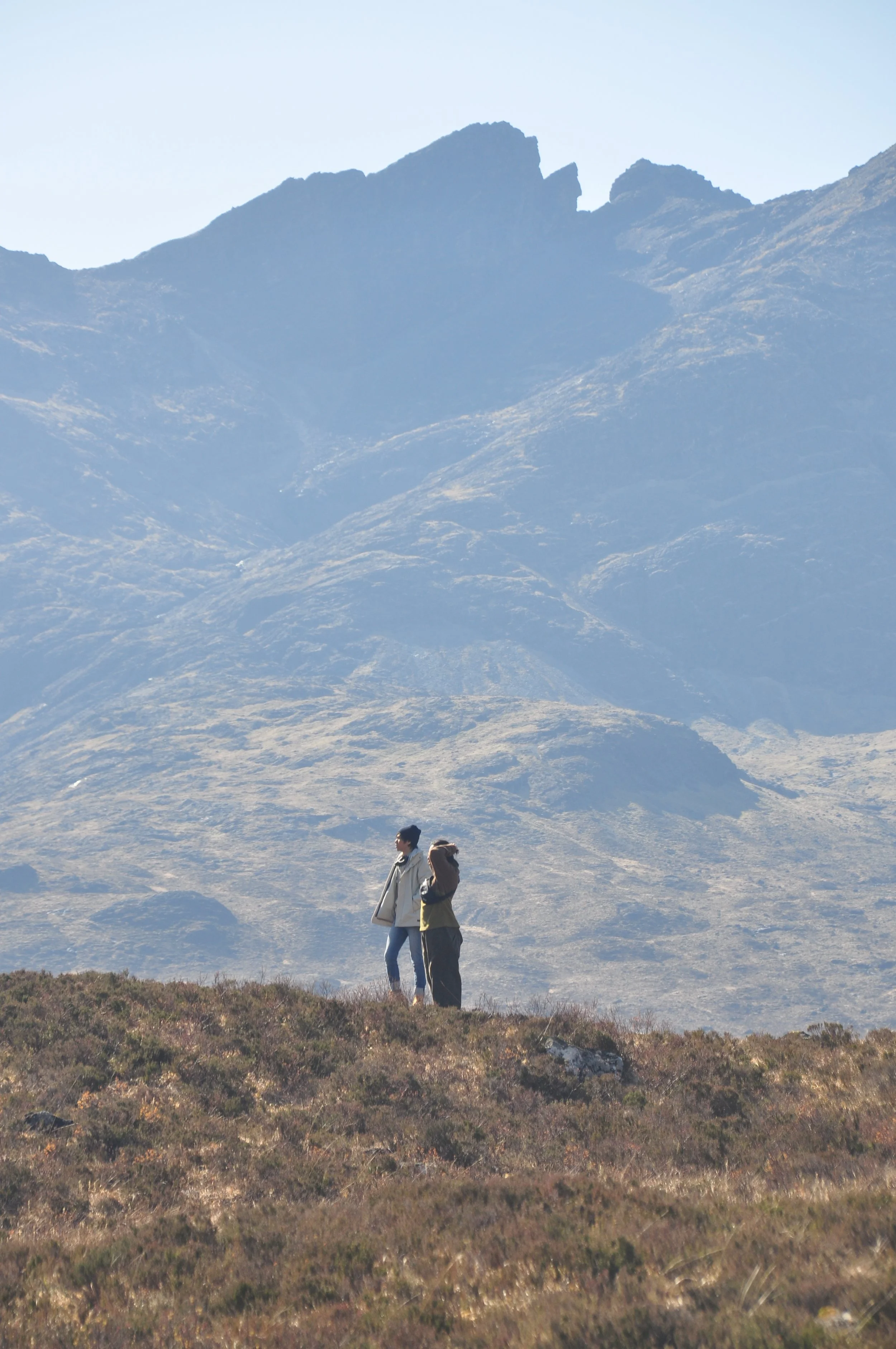 Two hikers wearing outdoor clothing standing in a mountainous landscape with rugged peaks in the background.