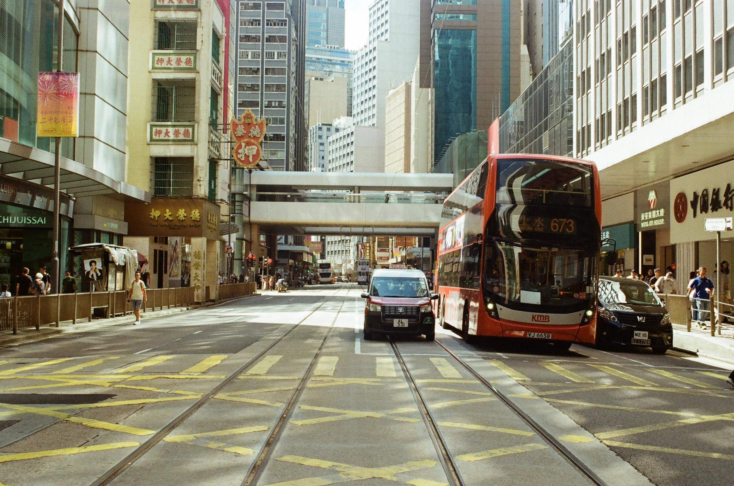 A busy city street with tram tracks, buses, cars, and pedestrians. Tall buildings line the street, with signs in Chinese characters and modern architecture.