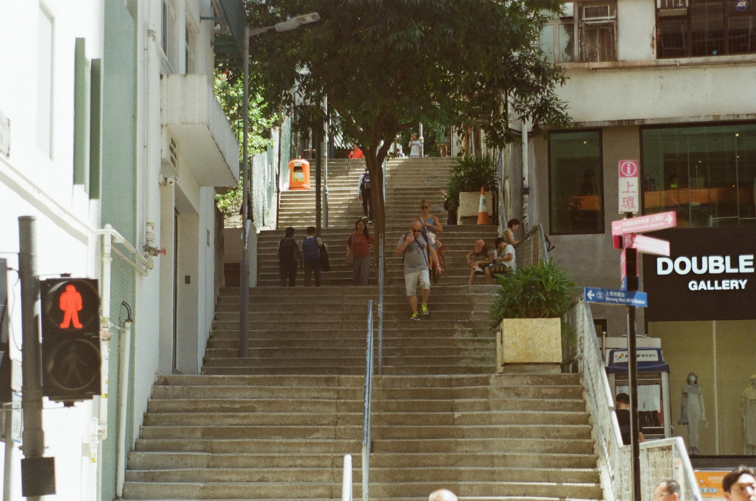 City sidewalk with wide staircase, several people walking up and down, some sitting on benches, a traffic light showing red walking symbol, and signs for Double Gallery and Sheung Wan MTR Station.