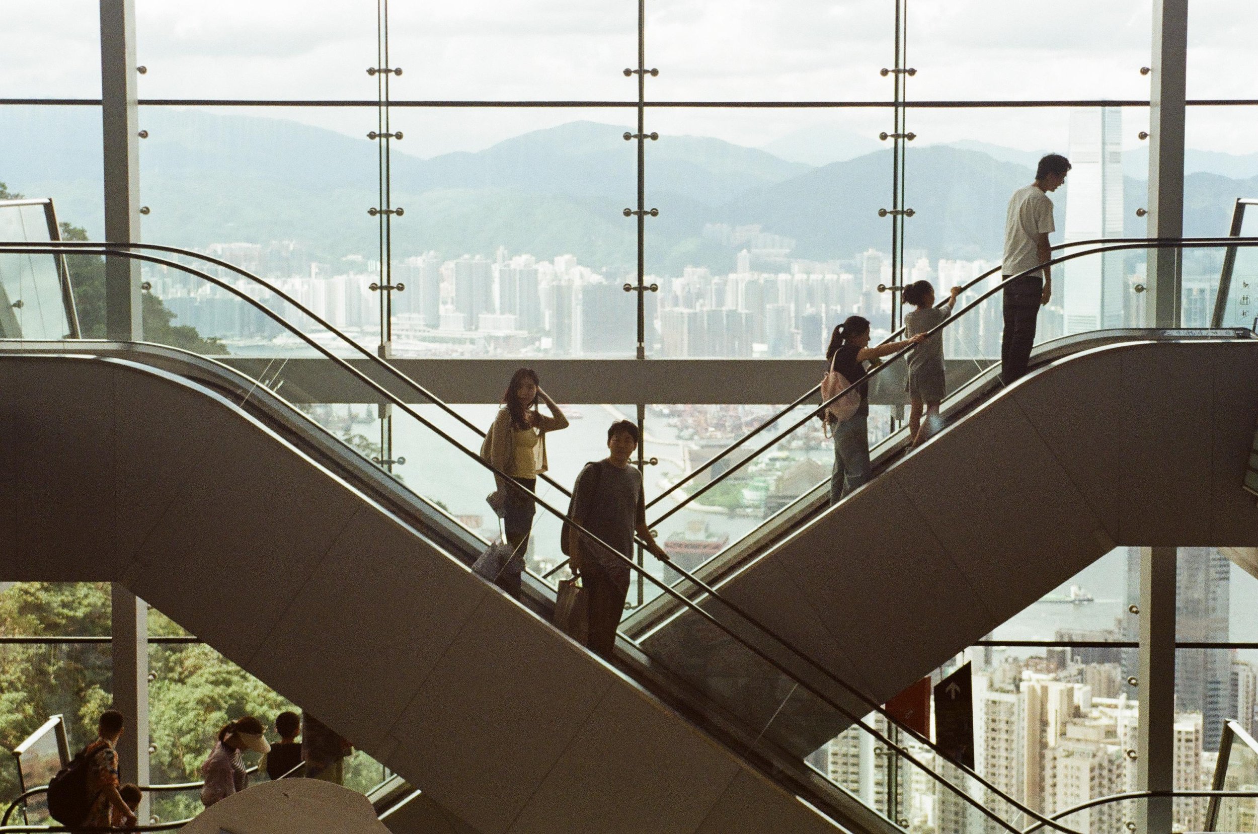 People descending an escalator inside a modern building with large glass windows and a city skyline in the background.