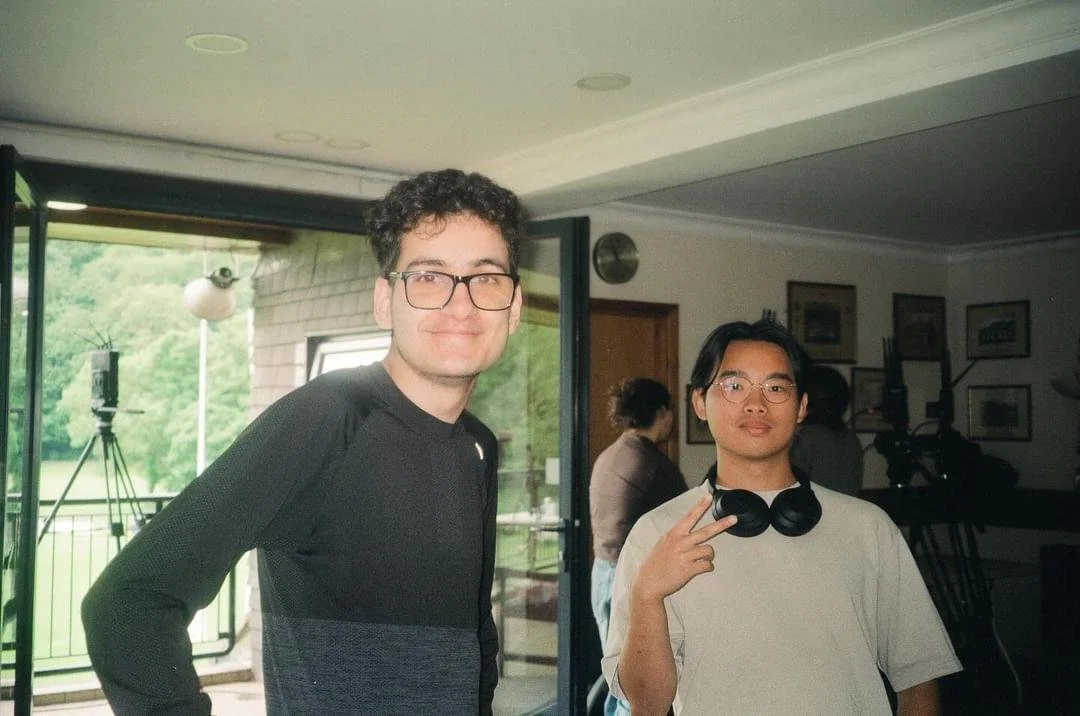 Two young men standing indoors, one making a peace sign with his fingers. The background shows a window, a balcony, and some framed pictures on the wall.