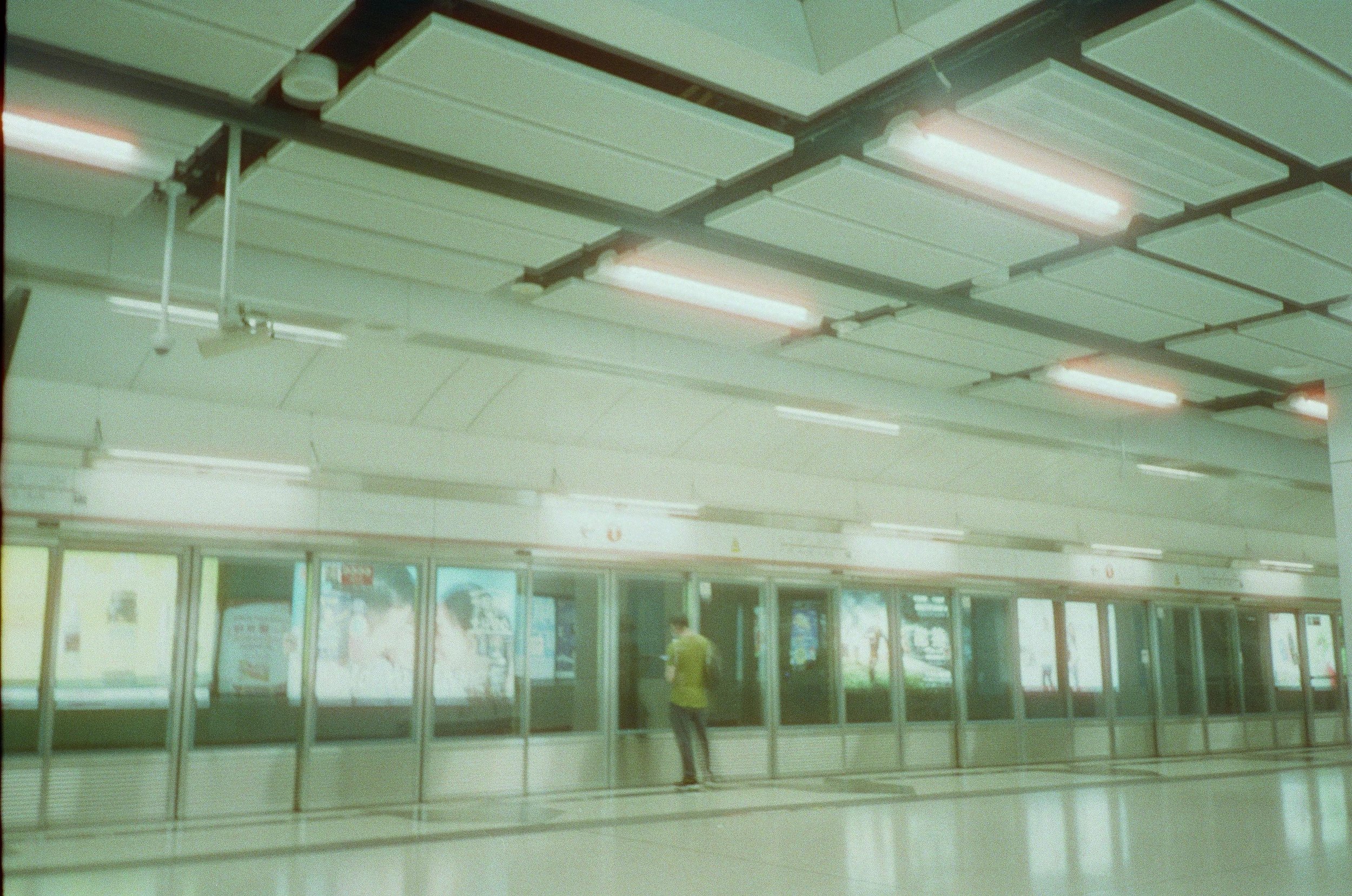 Empty subway station platform with a person waiting near the glass doors, fluorescent ceiling lights, and advertisements behind the glass doors.