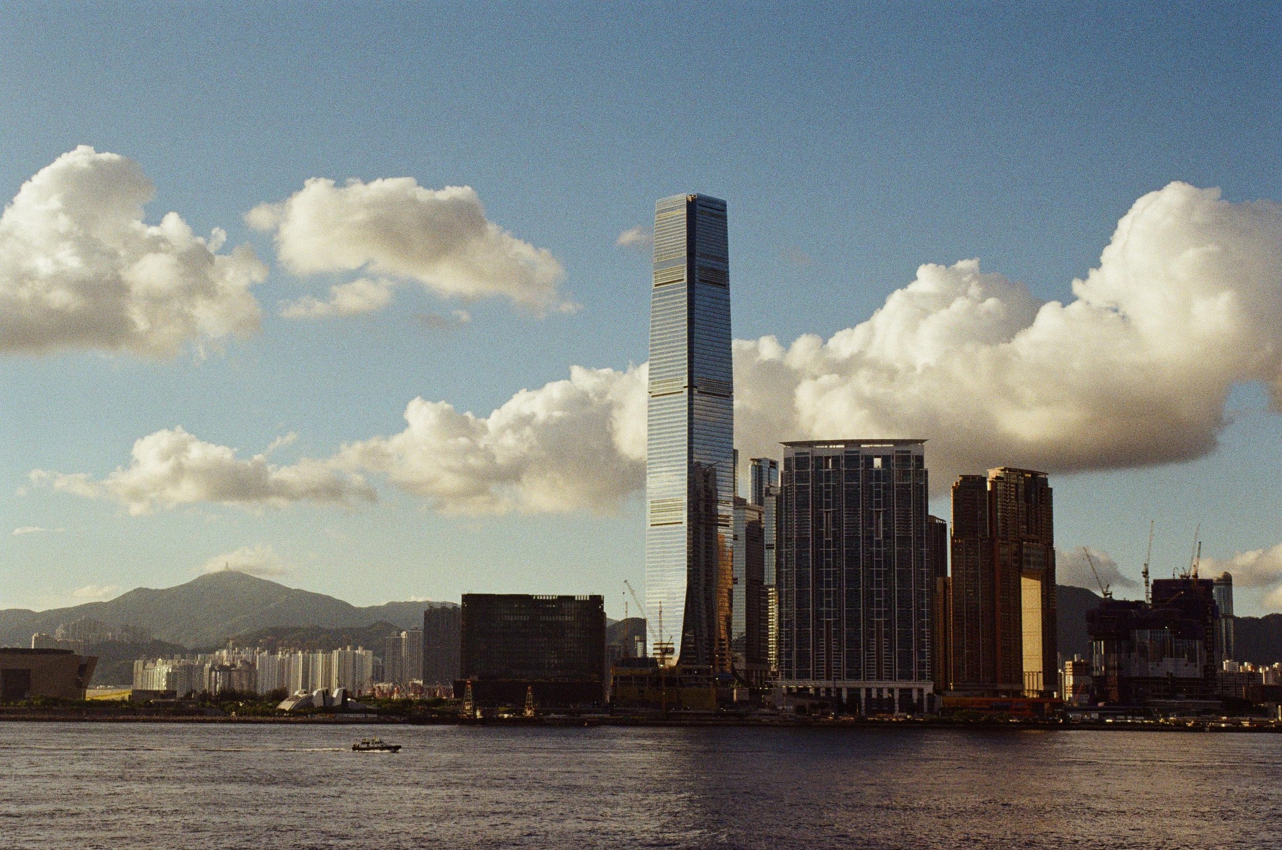 City skyline with tall modern skyscraper, high-rise buildings, and mountains in the distance, viewed across a body of water with a boat in the foreground.
