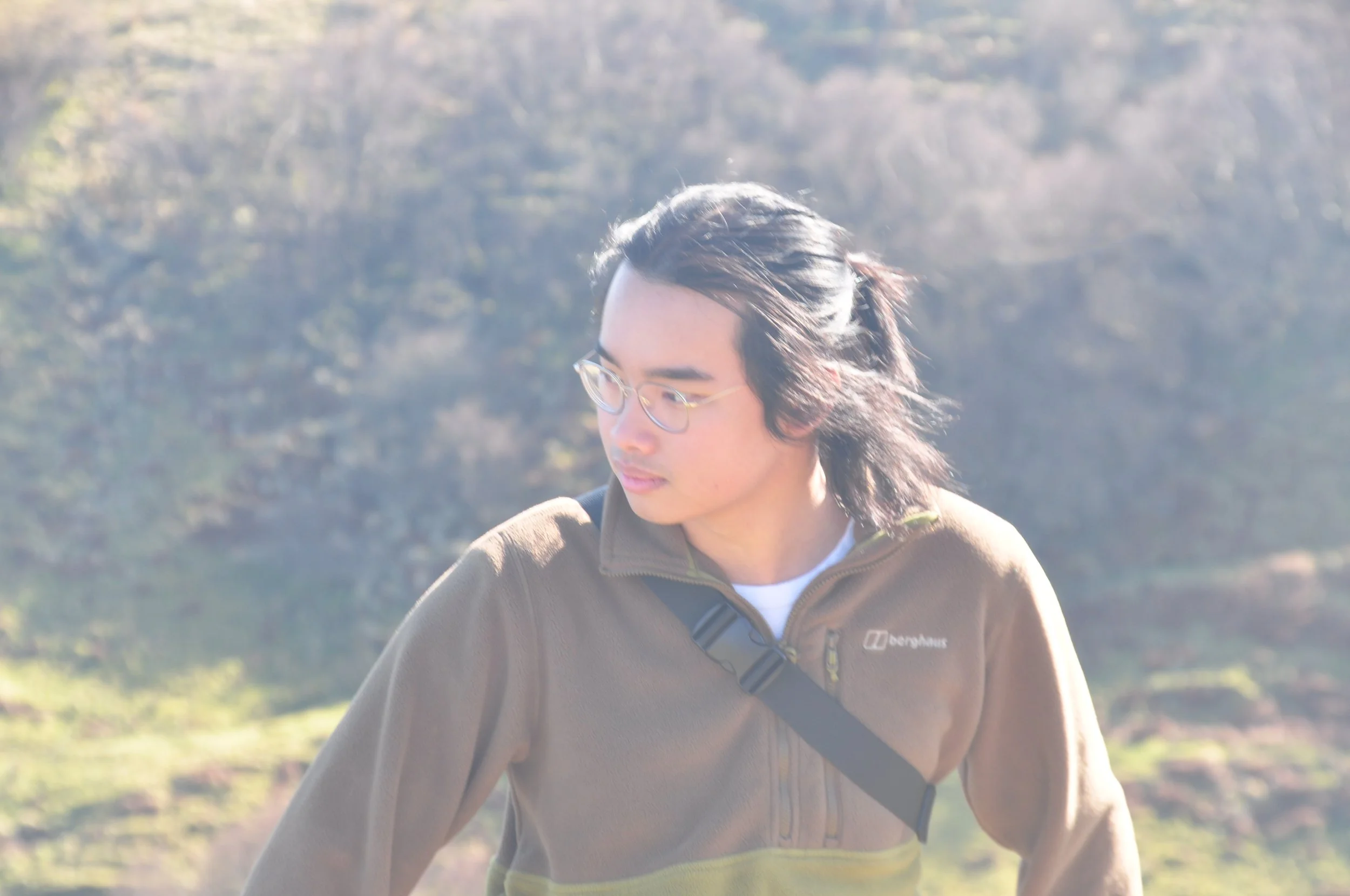 Young man outdoors wearing glasses and a brown jacket with a black strap over his chest, with a blurry natural landscape background.