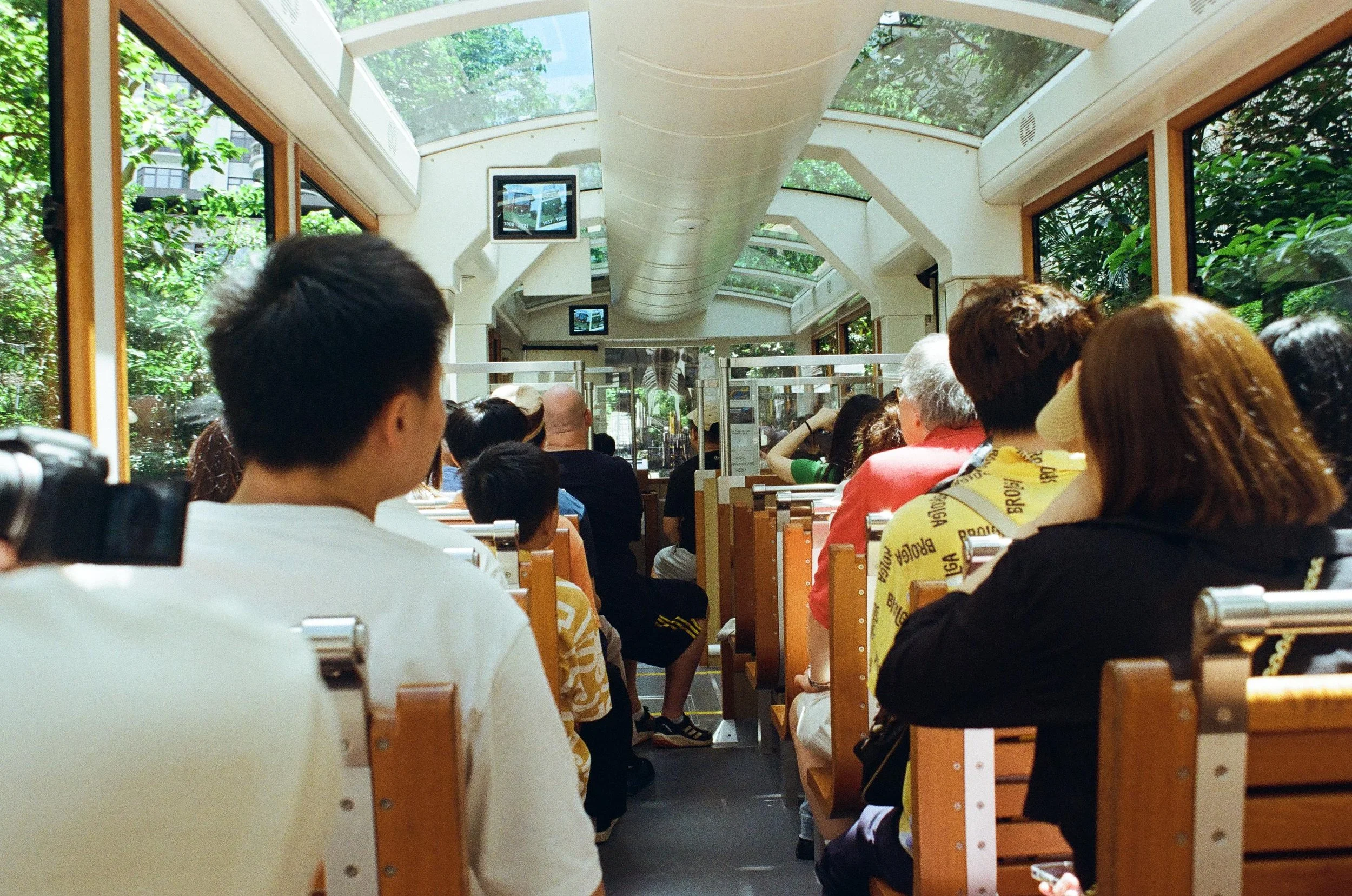 Inside a tram with many passengers seated and facing forward, with large windows revealing green trees outside, and screens hanging from the ceiling.
