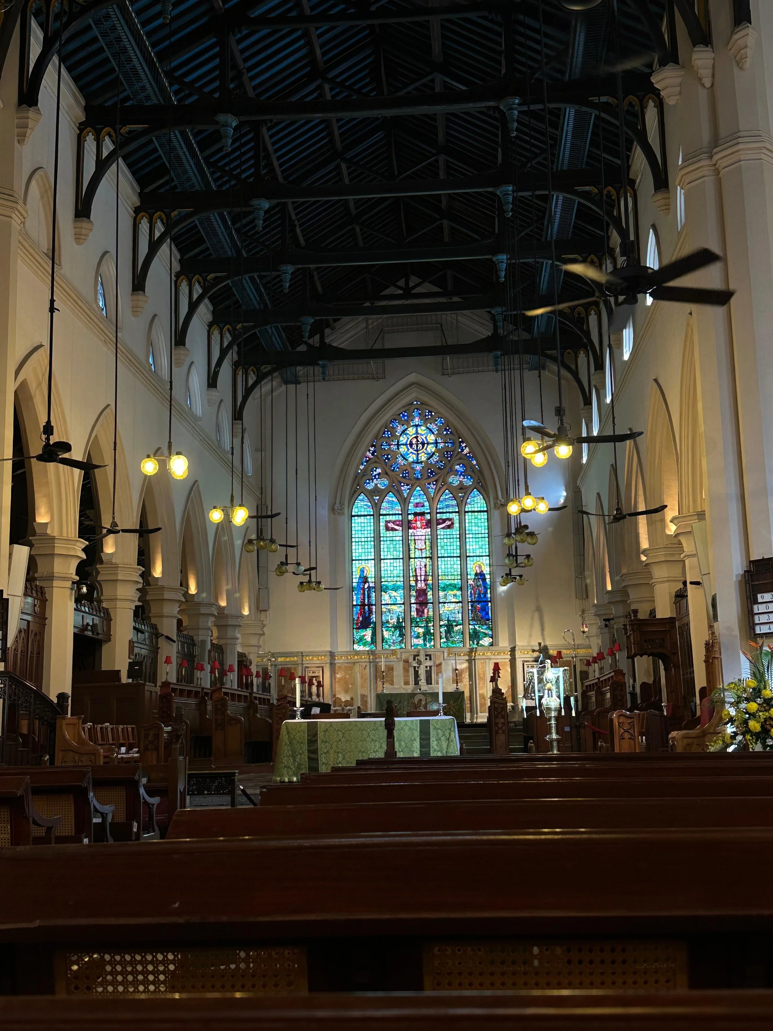 Interior of a church with wooden pews, stained glass windows depicting religious scenes, and an altar at the front. The ceiling is high with exposed dark beams, and there are hanging lights and ceiling fans.