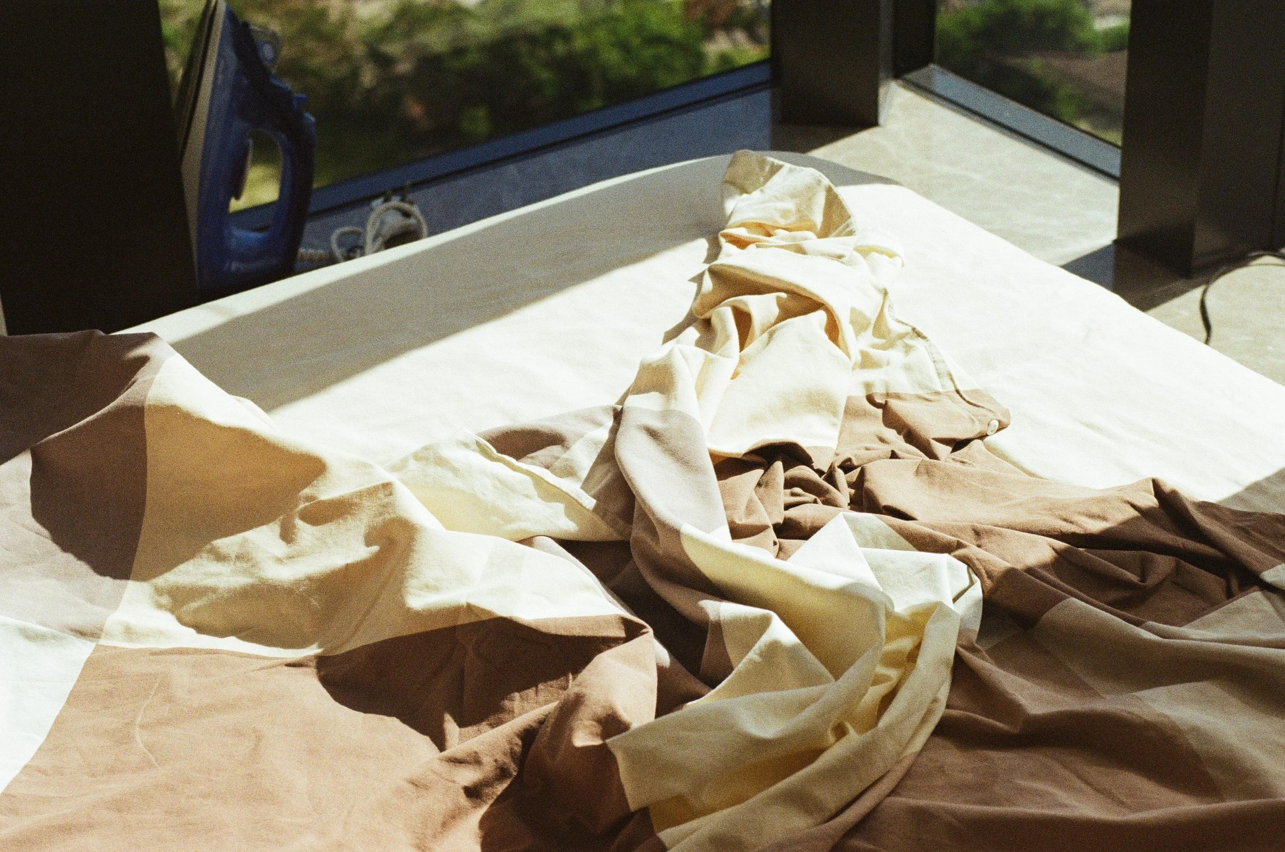 A crumpled bed with beige, cream, and brown sheets in a sunlit room with large windows.