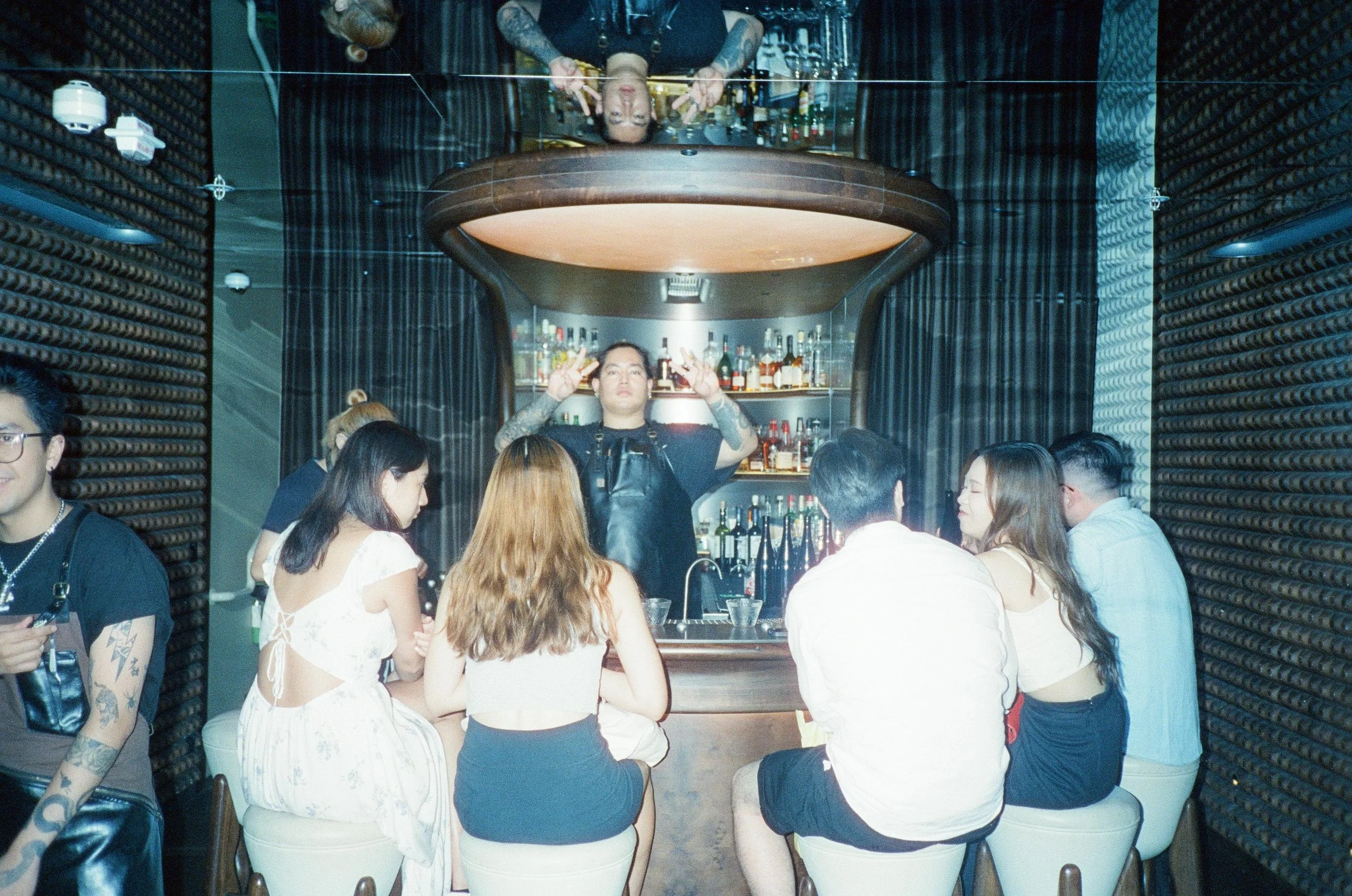 A bartender with tattoos making cocktails at a bar surrounded by a group of six diverse young adults seated on white and brown stools, in a modern bar with dark wood and black decor.