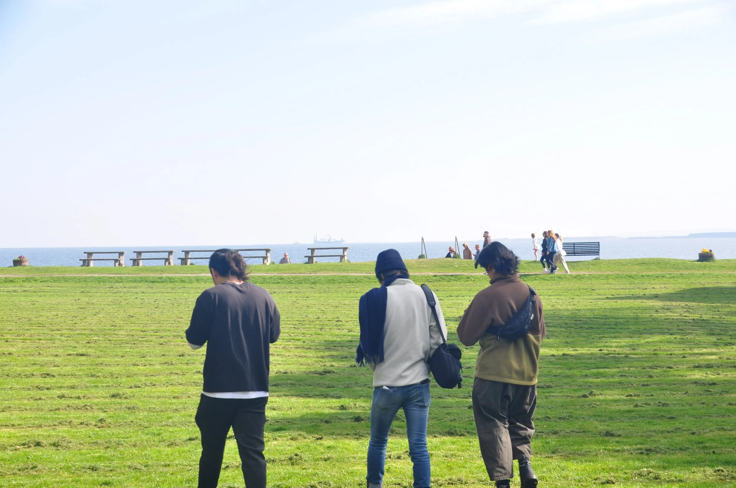 Three people in casual clothing standing on a grassy field near the water, with more people walking and benches in the background under clear skies.