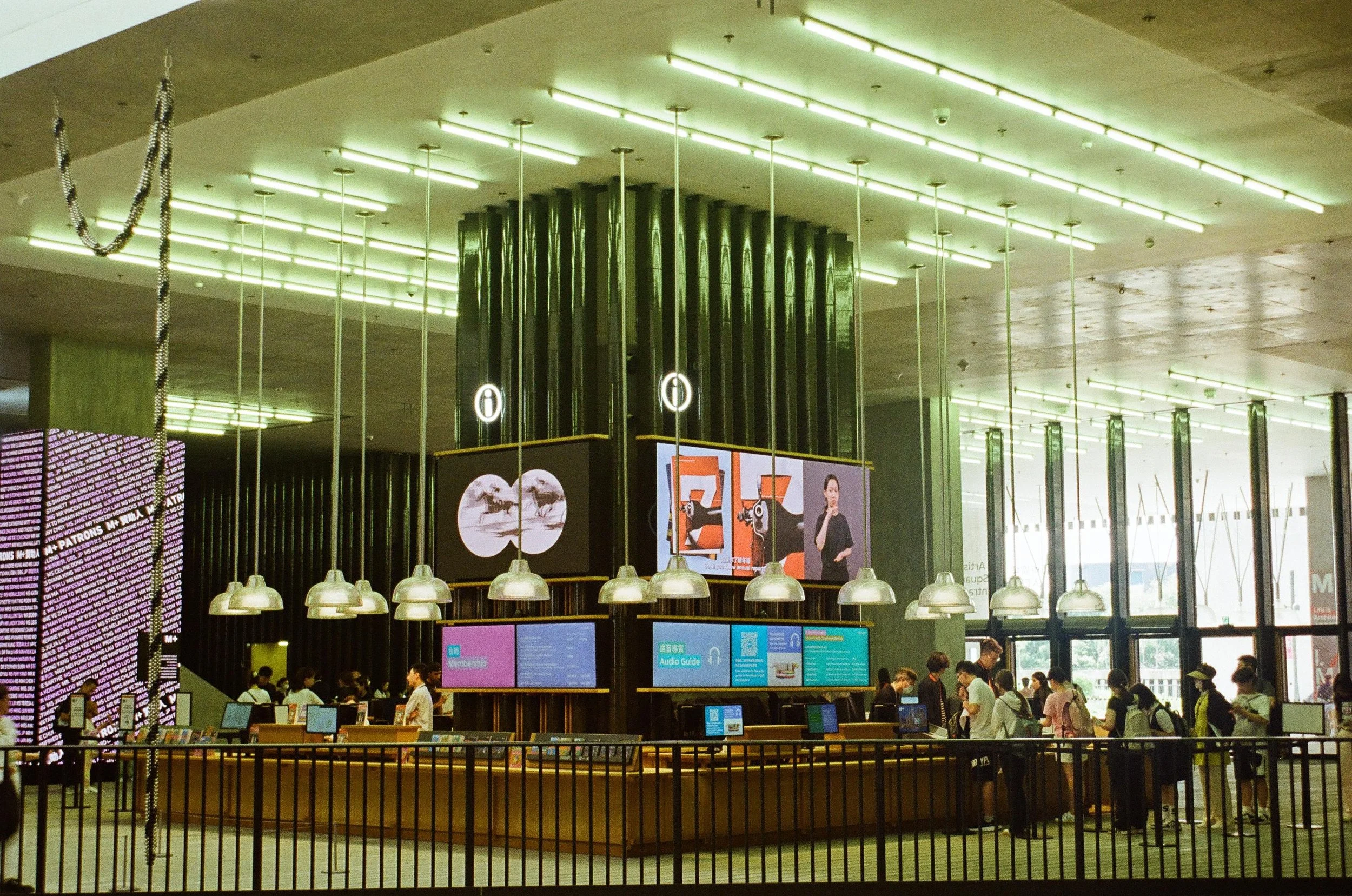 People line up at a transport ticket counter inside a modern building with large digital screens and high ceilings.