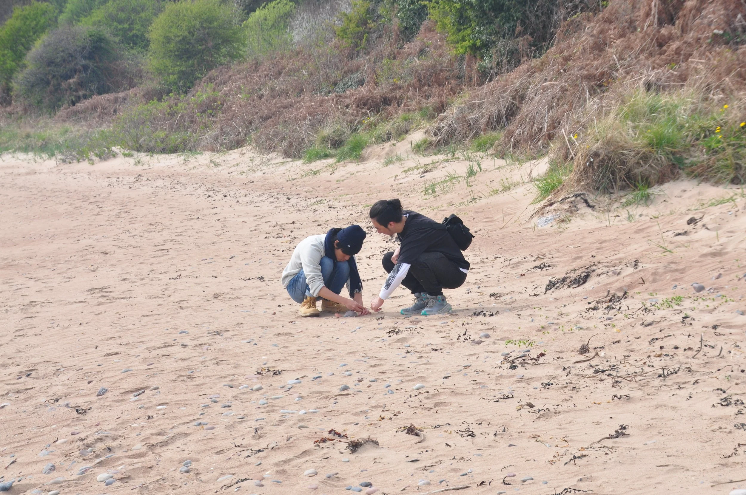 Two young men squatting on a sandy beach, examining something on the ground.