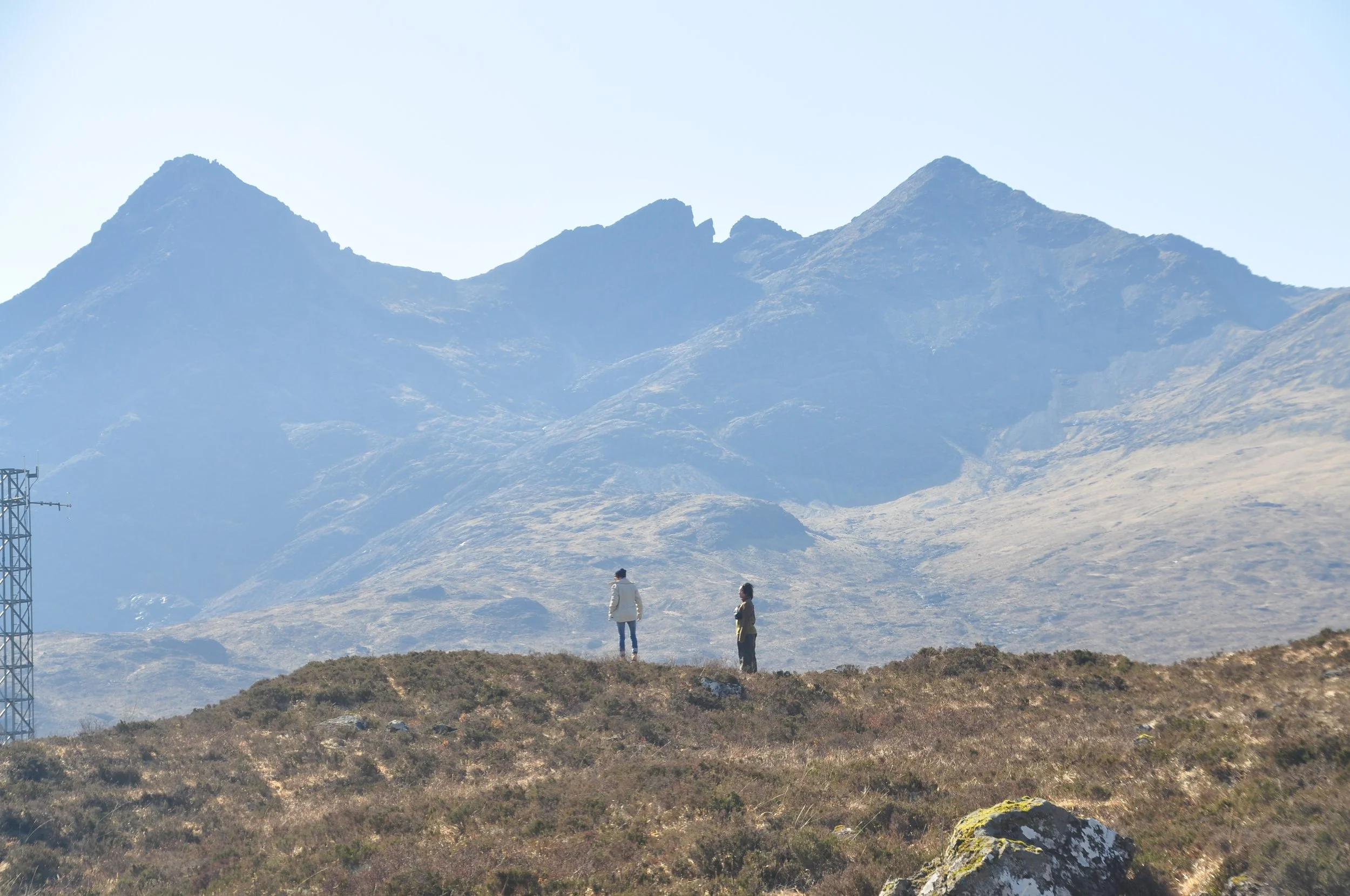 Two people standing on a grassy hill with mountain range in the background.