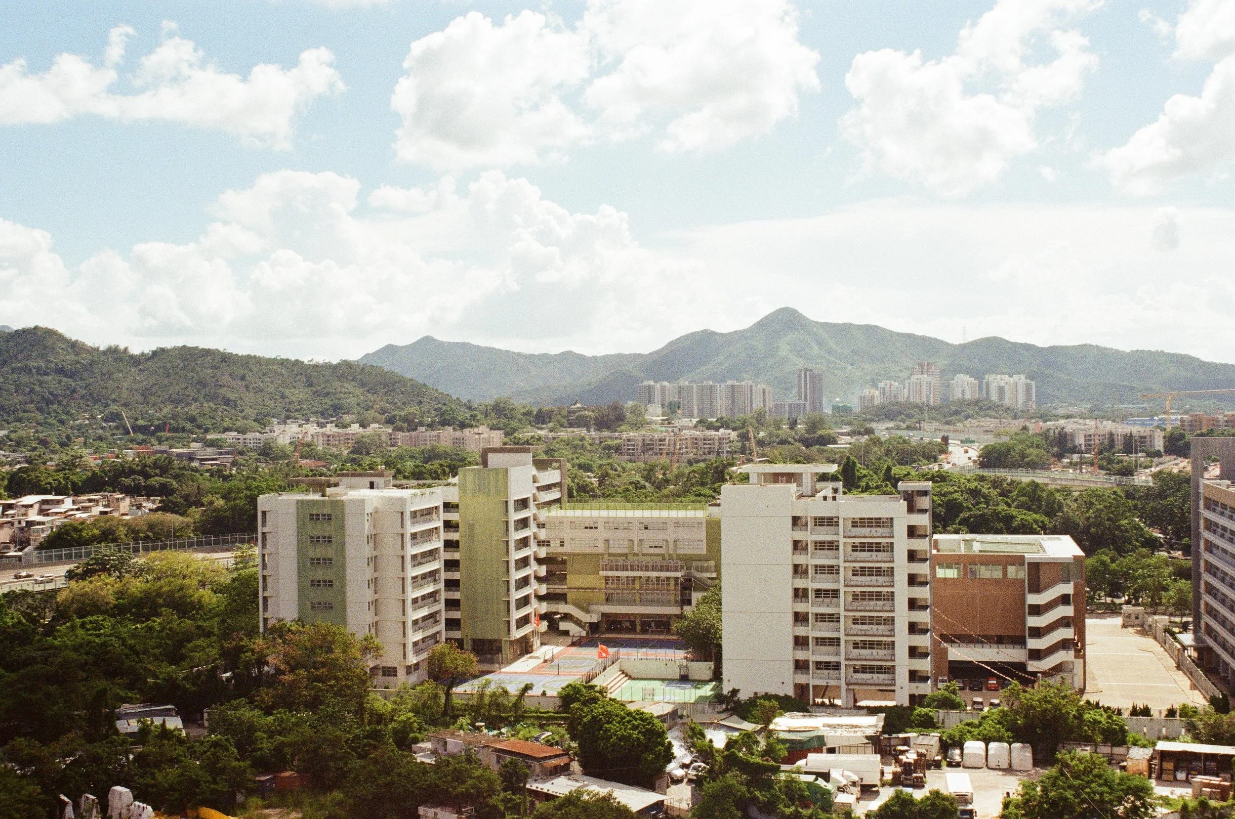 A cityscape with several high-rise buildings, green trees, mountains in the background, and a partly cloudy sky.
