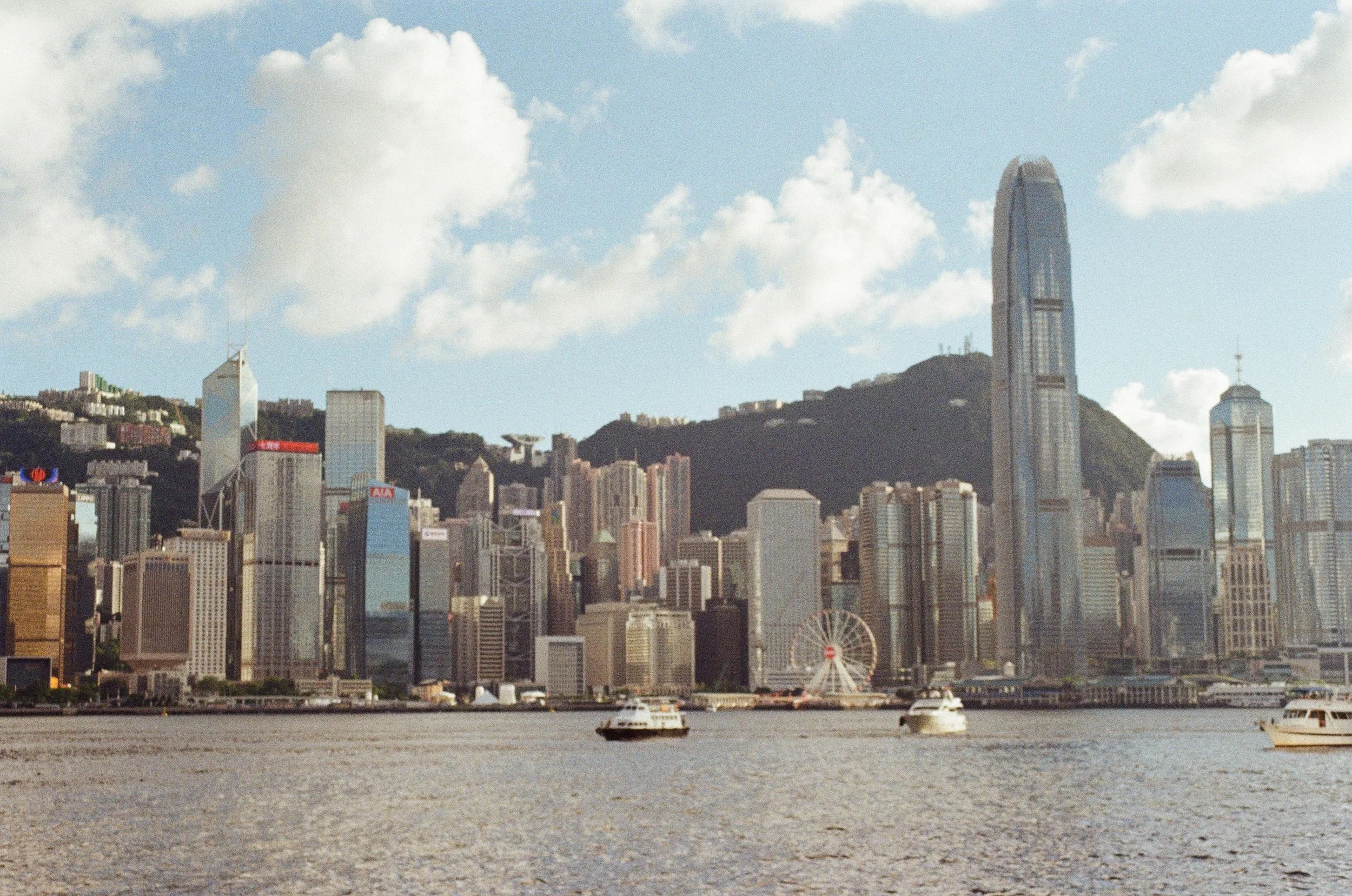 City skyline of Hong Kong with tall skyscrapers, a ferris wheel, boats on water, and mountains in the background under a partly cloudy sky.
