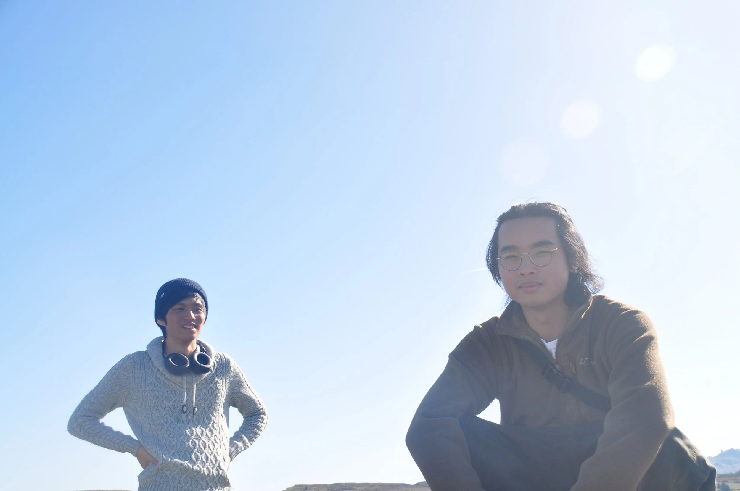 Two young men outdoors under a clear blue sky, one sitting in the foreground and the other standing in the background, both smiling.