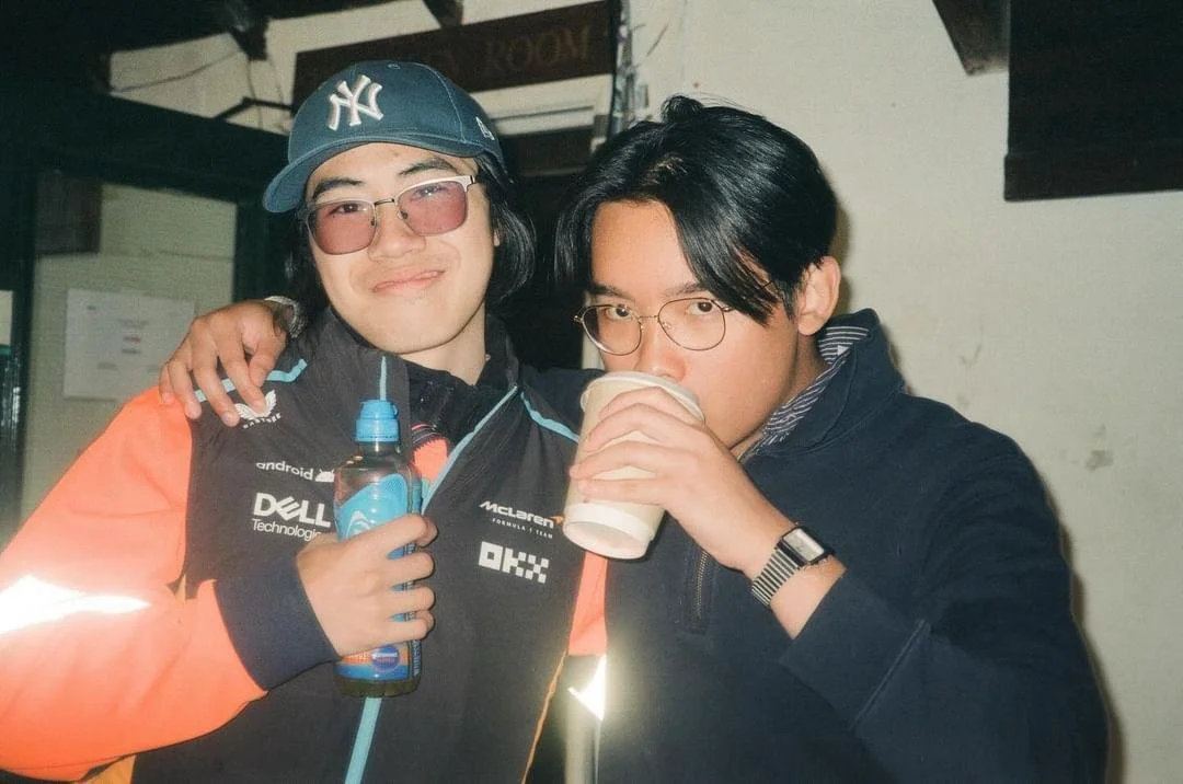 Two young men at a social gathering, one wearing a New York Yankees cap and casual glasses, the other wearing glasses and drinking from a paper cup, with the first person holding a water bottle.
