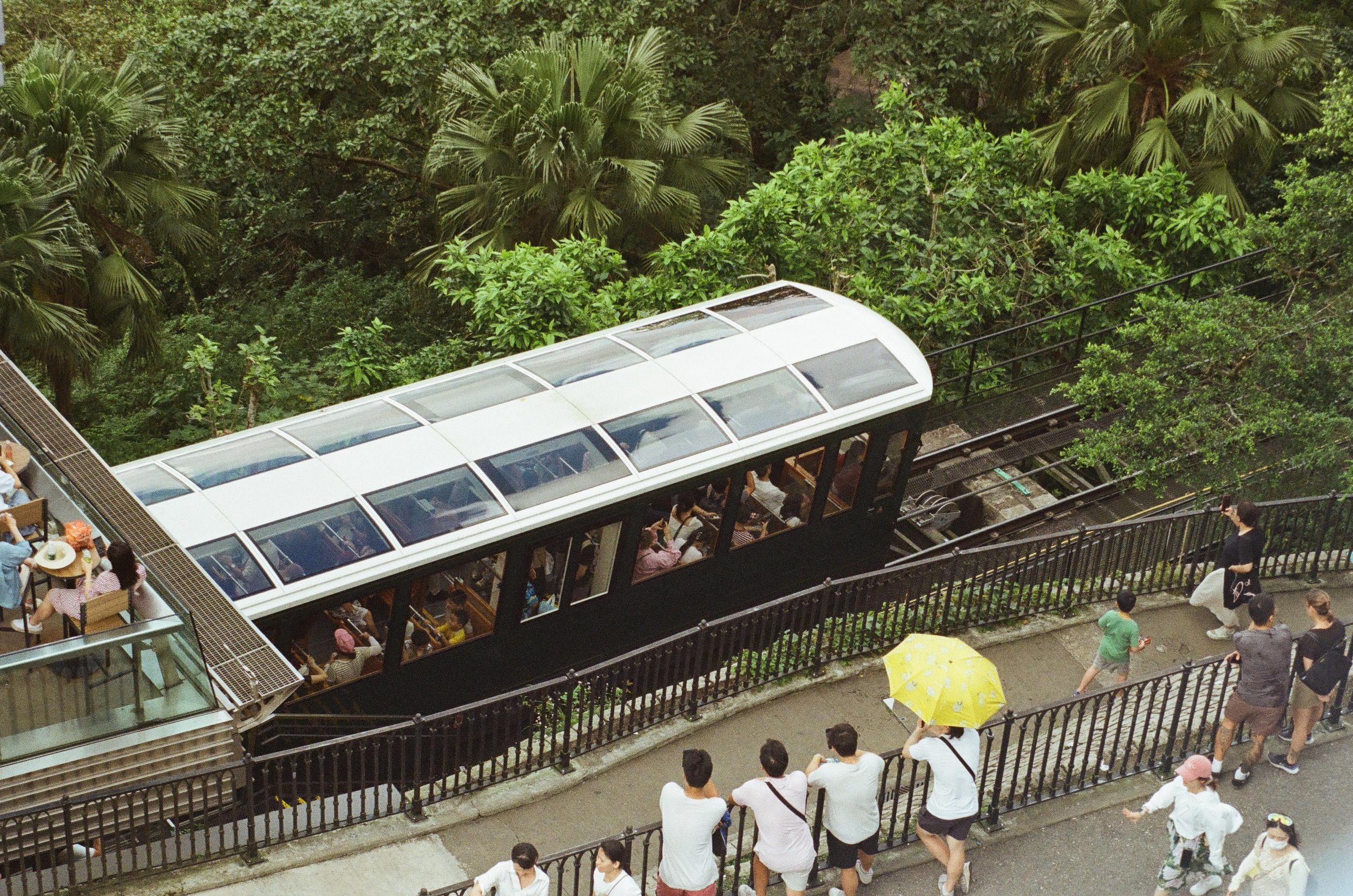 People riding an aerial tramway through a lush green forest, with onlookers standing by a fence and a person holding a yellow umbrella.