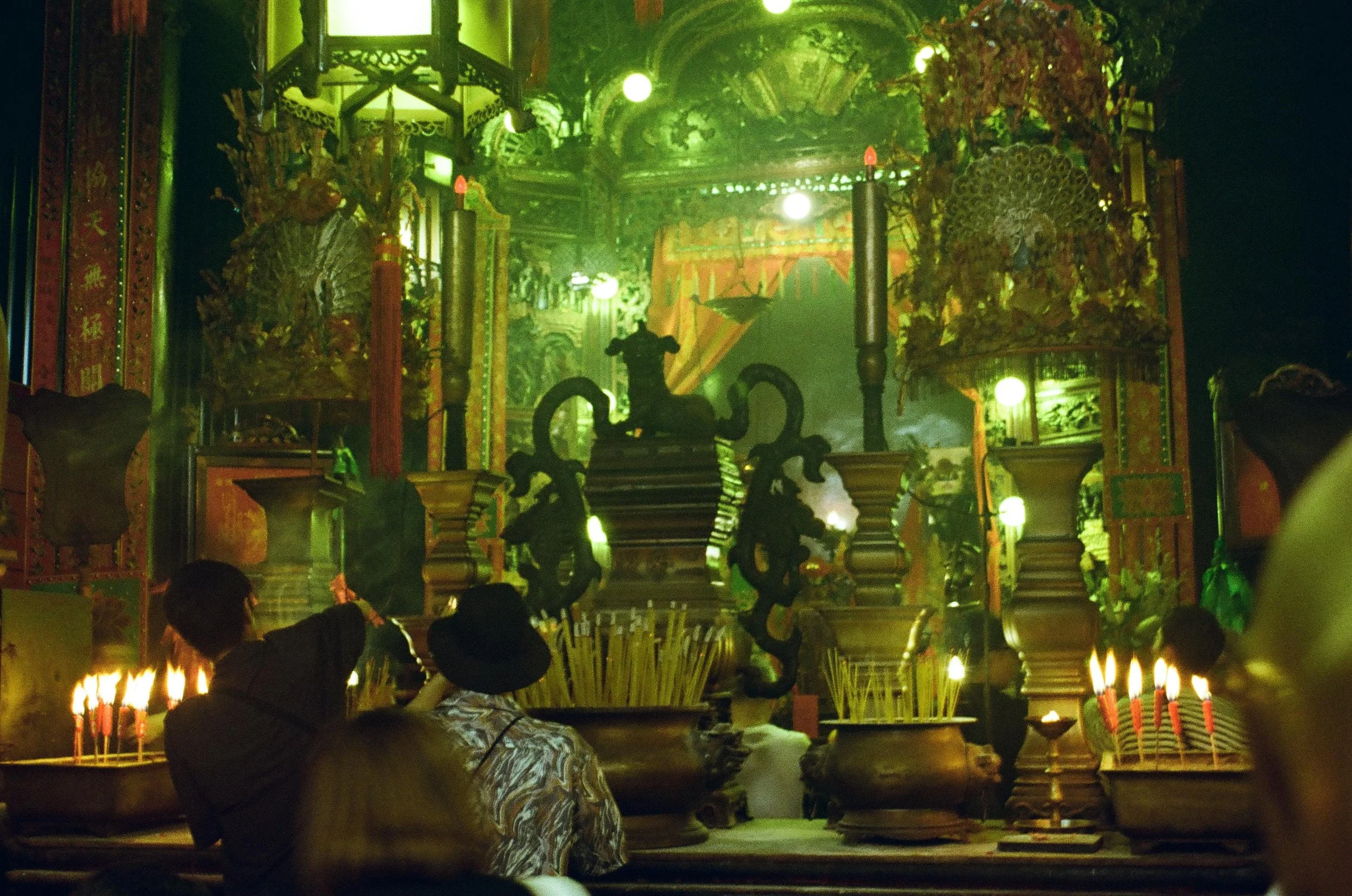 People praying inside a richly decorated Chinese temple with incense burners and illuminated hanging lanterns with green lighting.