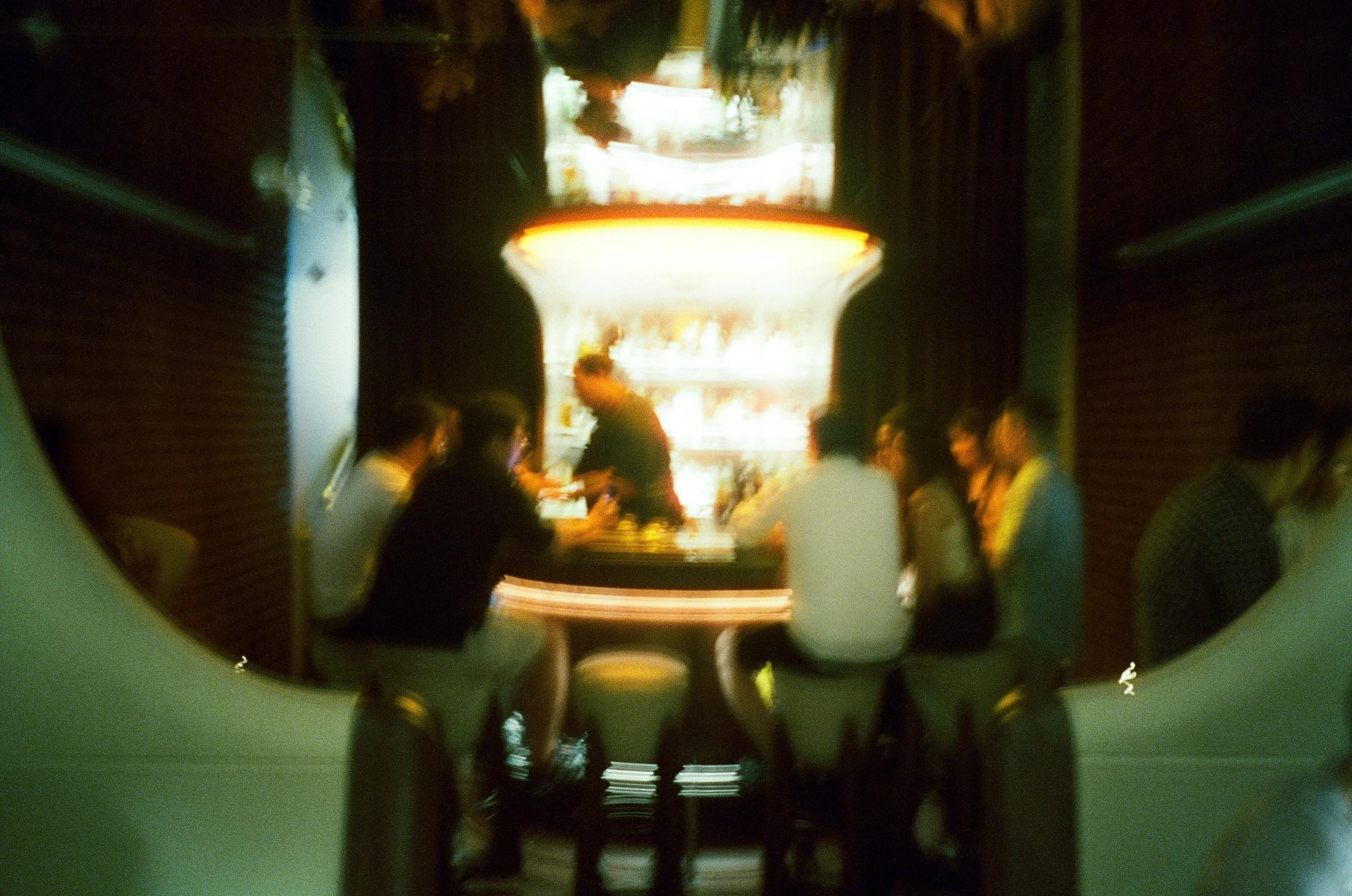 Blurred photo of a group of people sitting around a bar counter in a dimly lit setting.