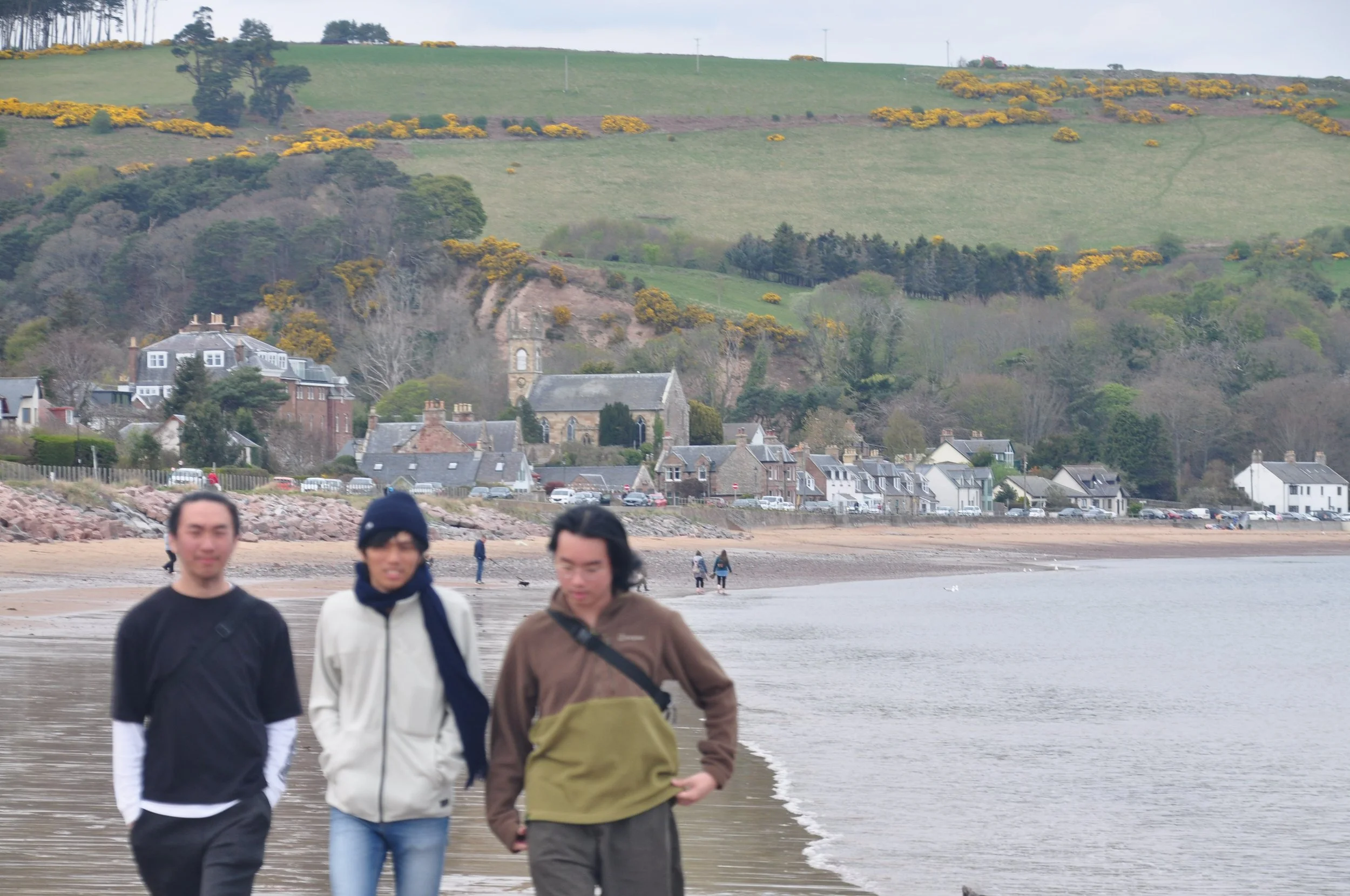 Three people walking along a beach with a village and green hills in the background.