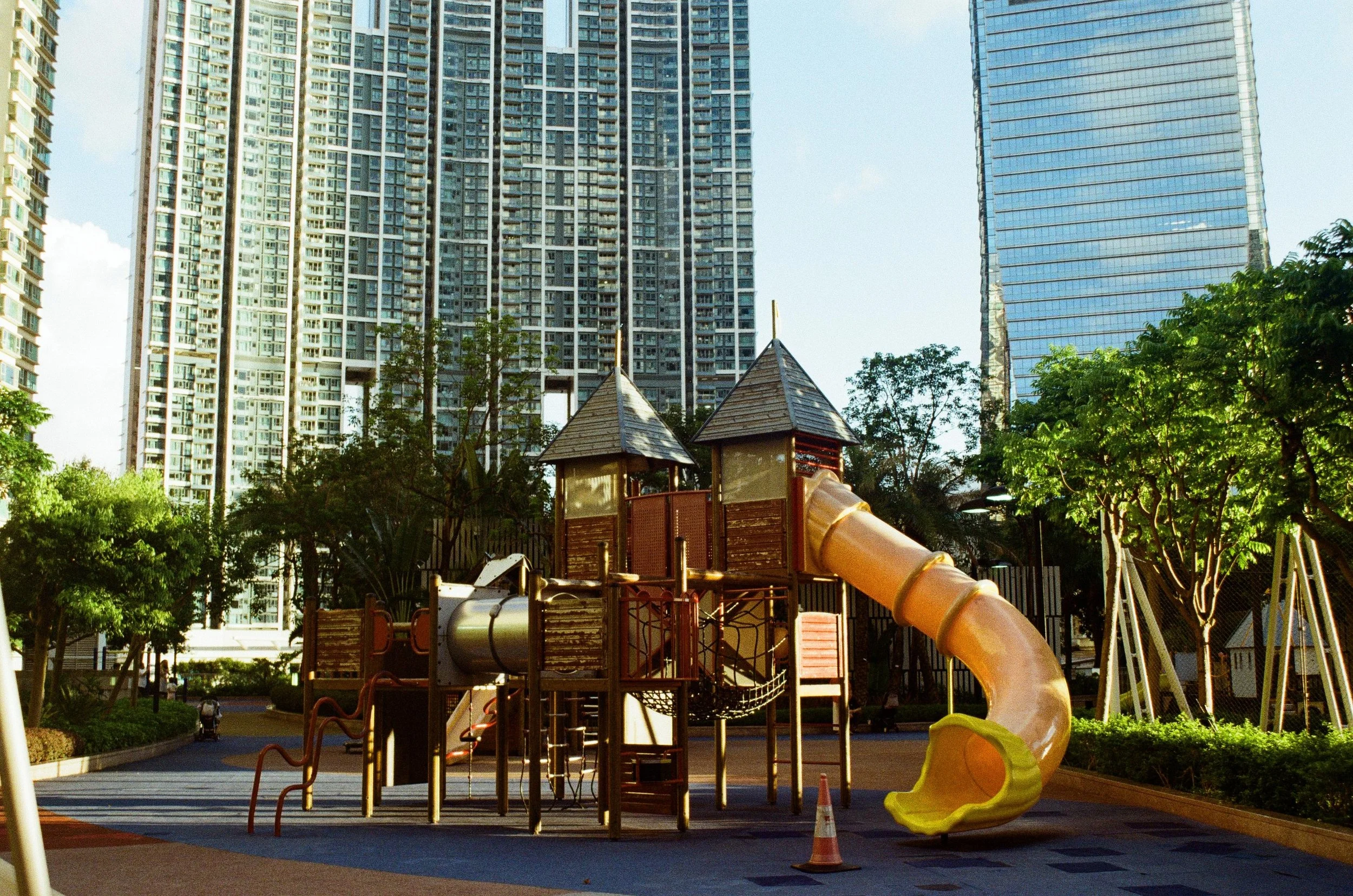 Empty children's playground with slide and climbing structures, surrounded by tall modern skyscrapers and trees.