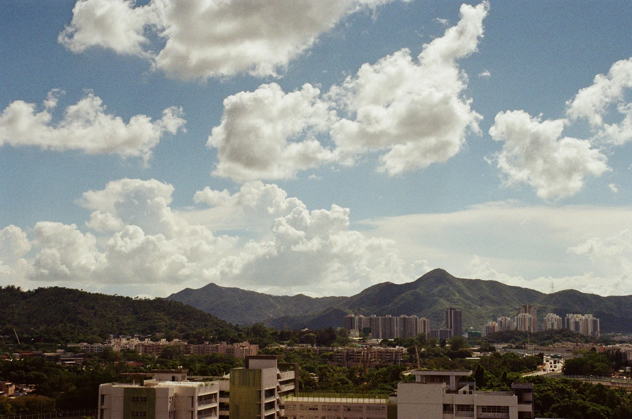 Cityscape with buildings and green mountains under a partly cloudy sky.